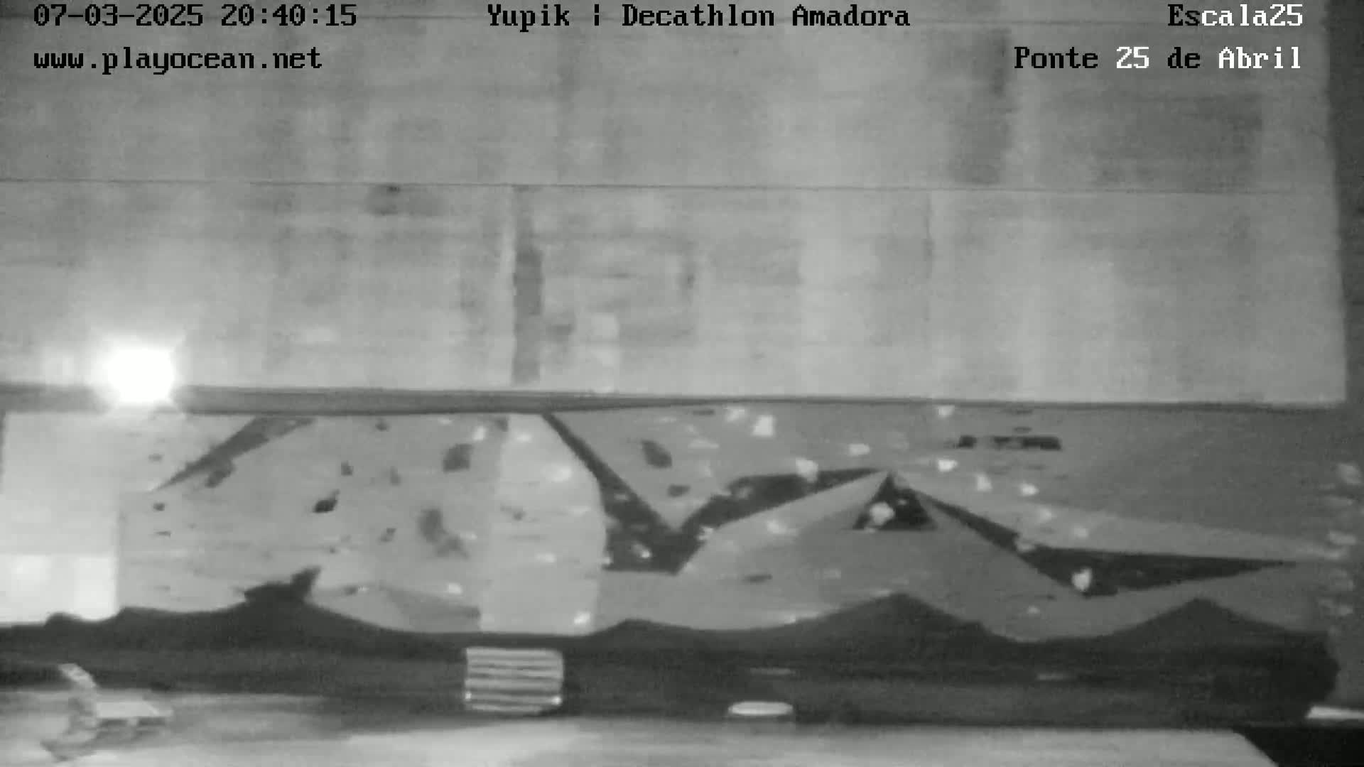 A nighttime, grayscale image shows a wall with a climbing structure, illuminated by a bright light source.