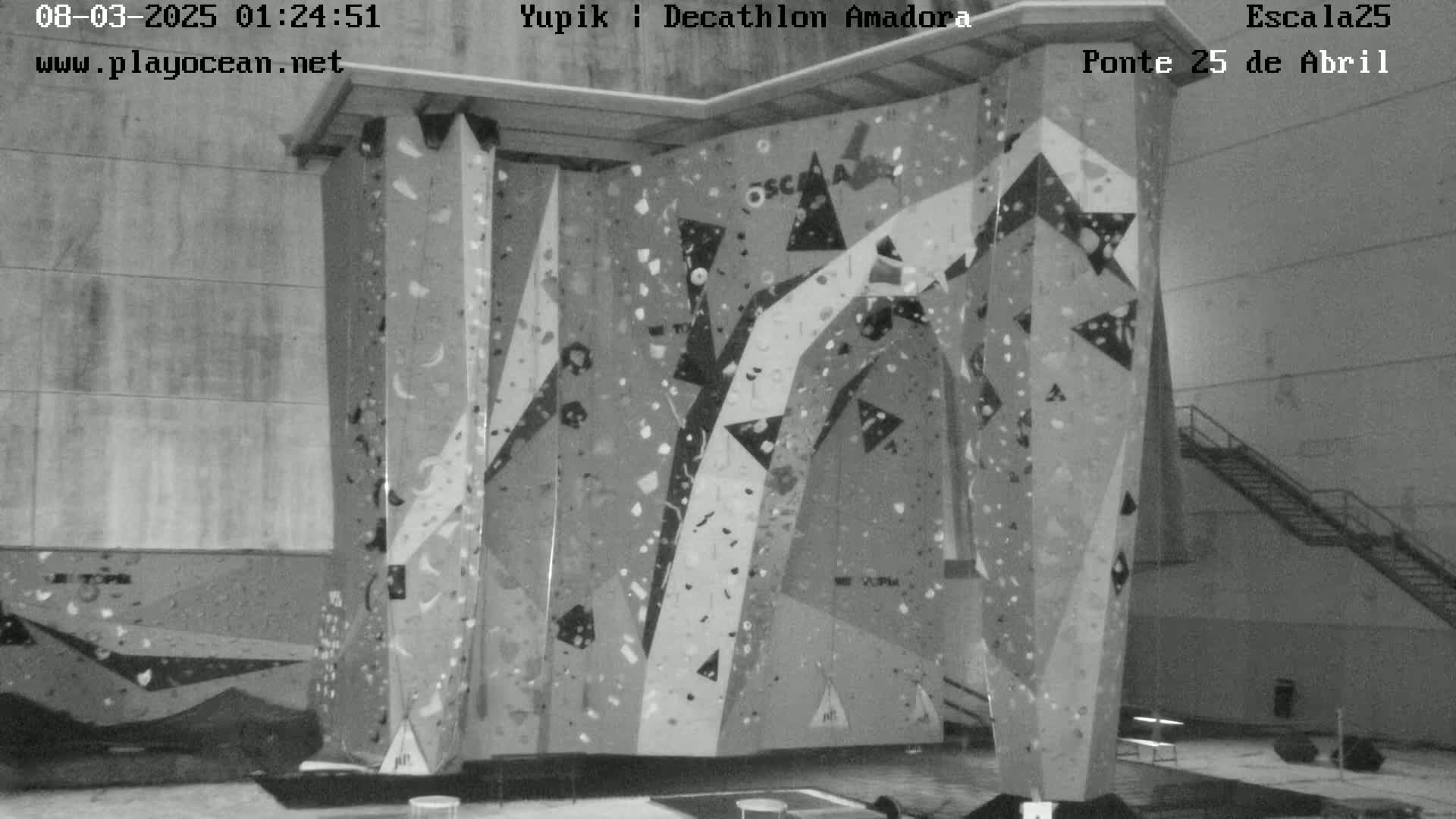 A grayscale indoor image shows a large, multi-sectioned climbing wall with various holds and patterns.