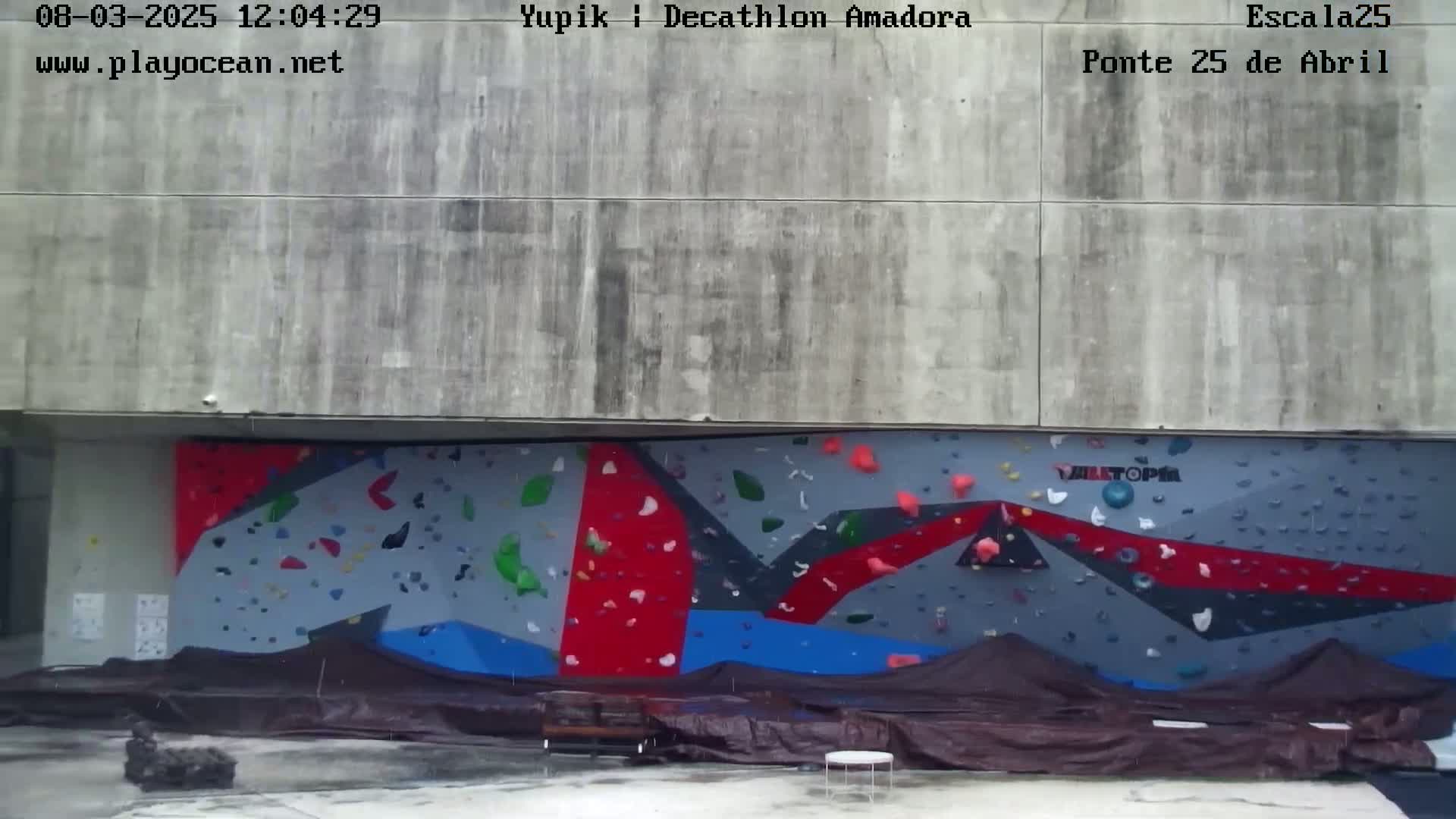 A large outdoor climbing wall with red, blue, and gray sections is partially covered by brown tarp in rainy weather.