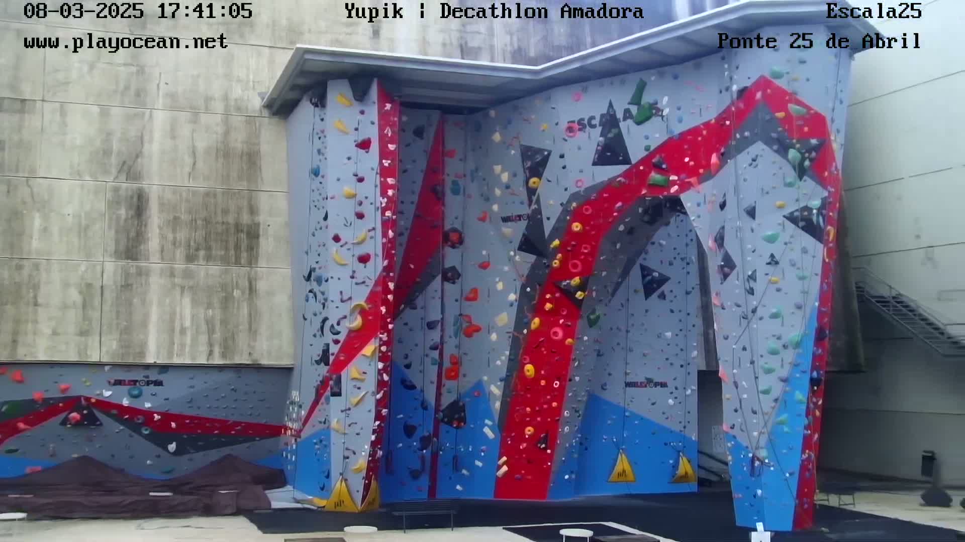 A large, colorful outdoor climbing wall stands against a light gray building on an overcast day.