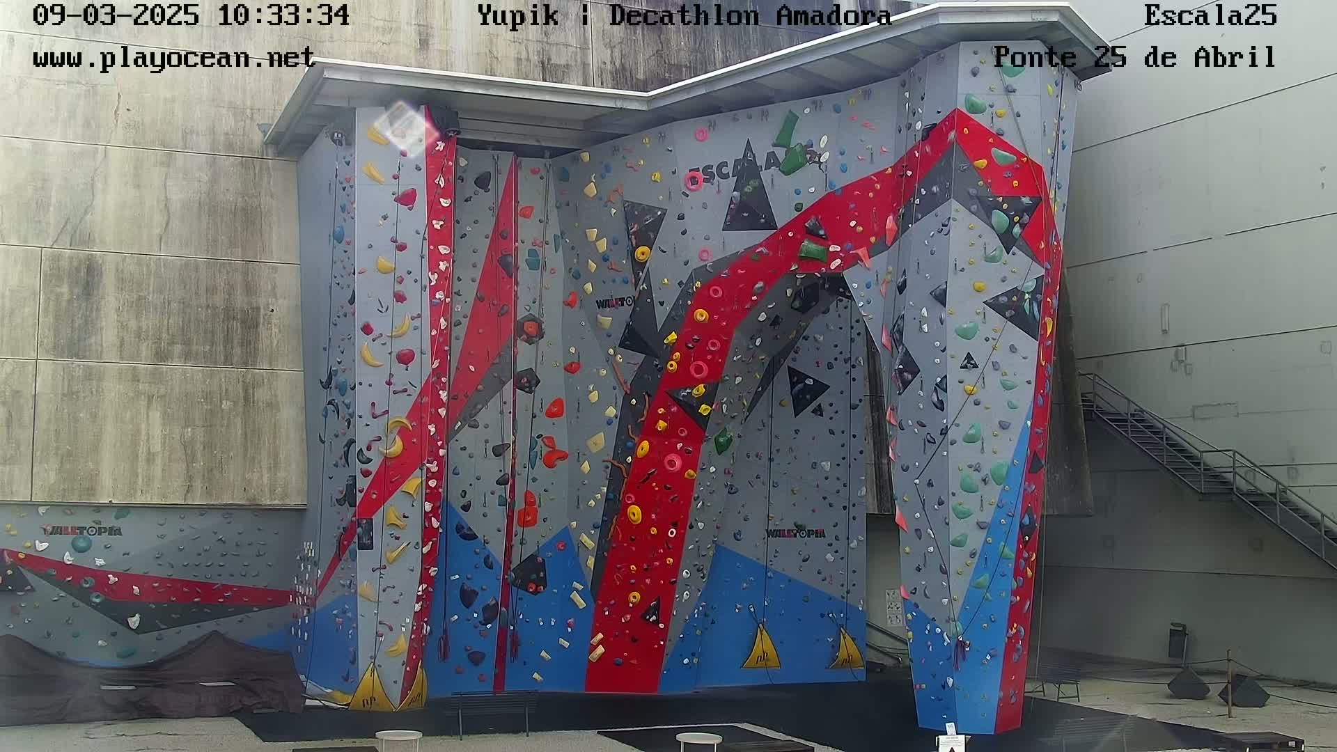 An outdoor, brightly colored climbing wall with various holds stands against a light gray building next to a metal staircase.