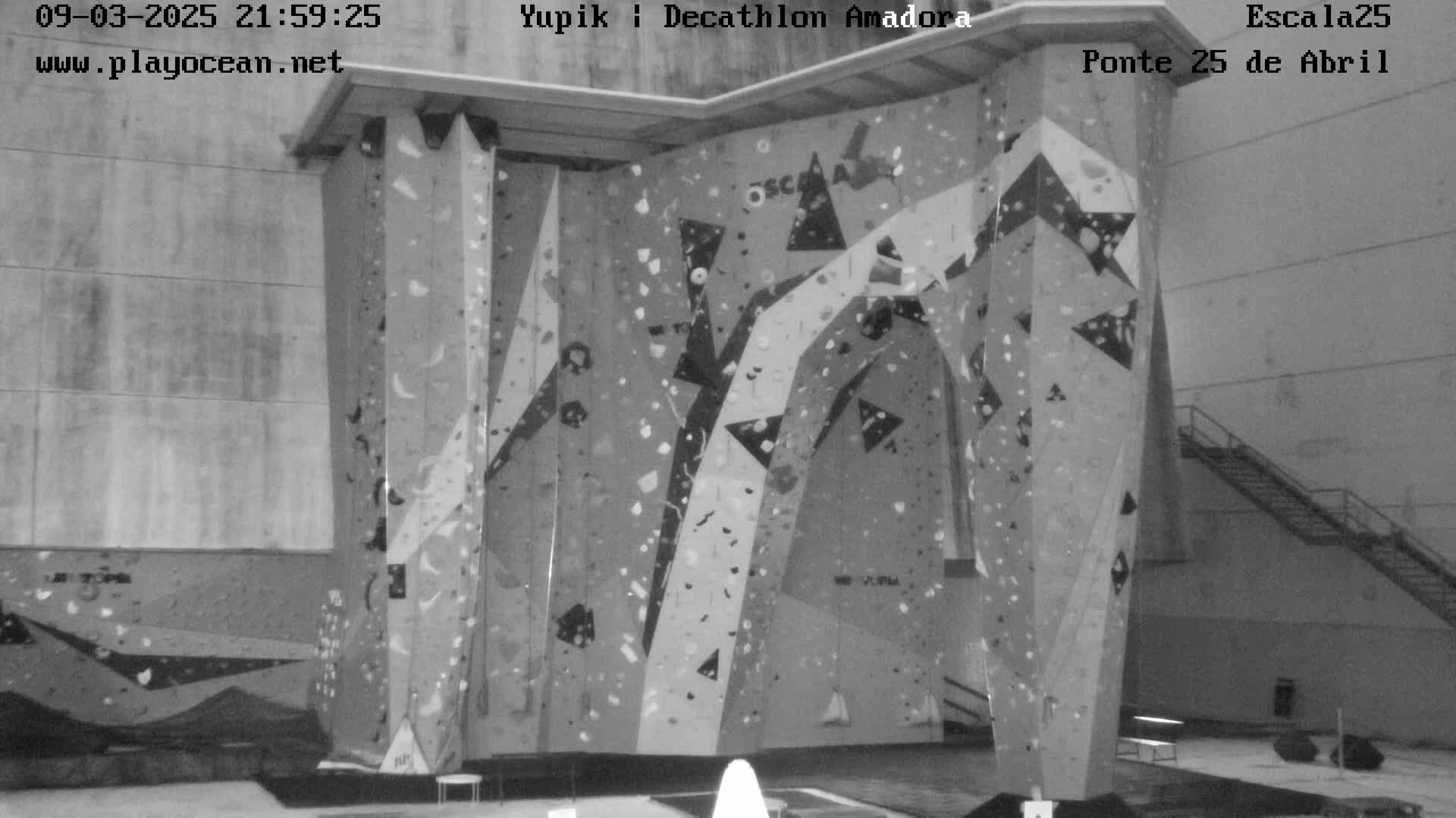A black and white indoor image shows a large climbing wall with various holds and patterns.