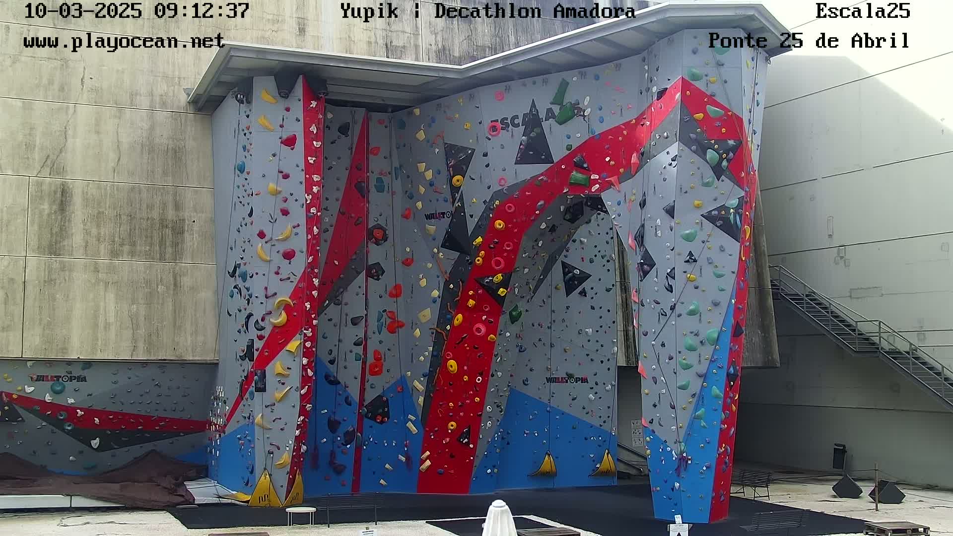 An outdoor, brightly colored climbing wall with various holds stands against a light gray building on a partly cloudy day.