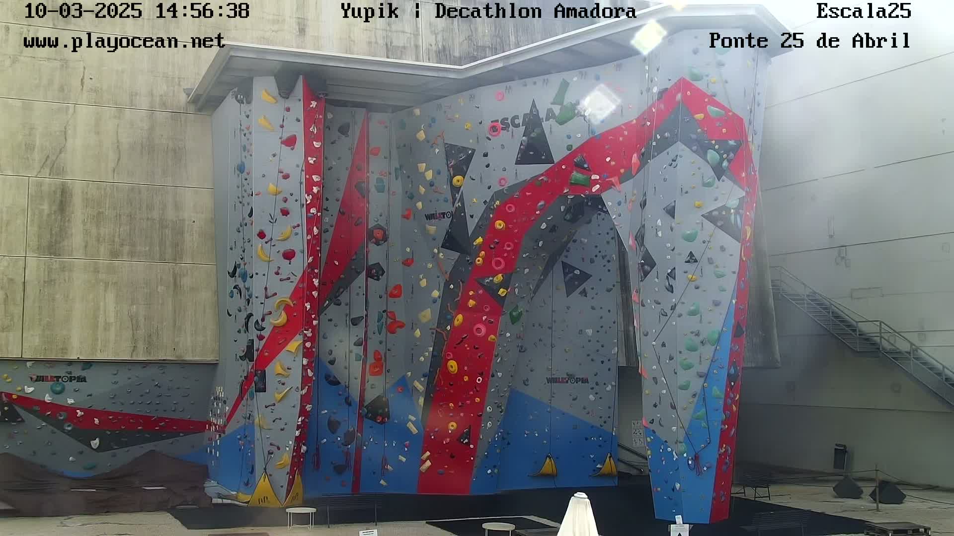 A large indoor climbing wall with various colored holds is situated against a light gray building on a sunny day.