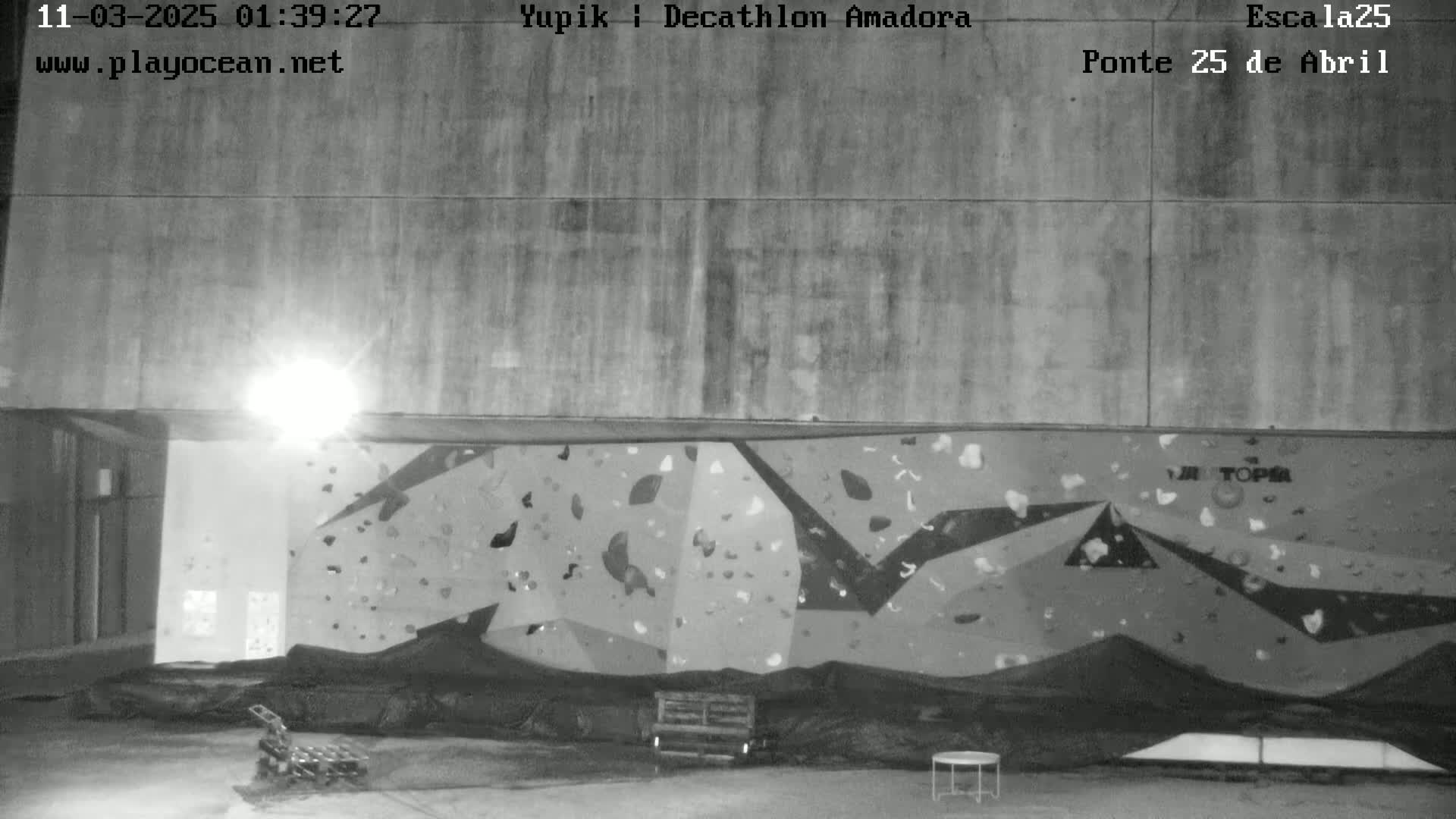 A nighttime, black and white image shows an indoor climbing wall with various holds affixed to its surface.