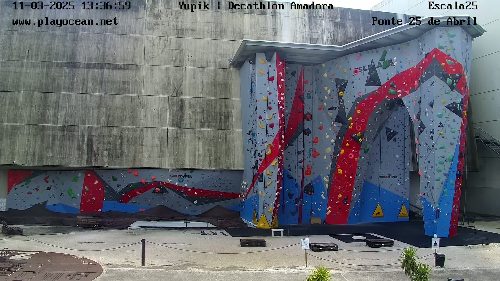 An outdoor, brightly colored climbing wall is situated against a large gray building on a sunny day.