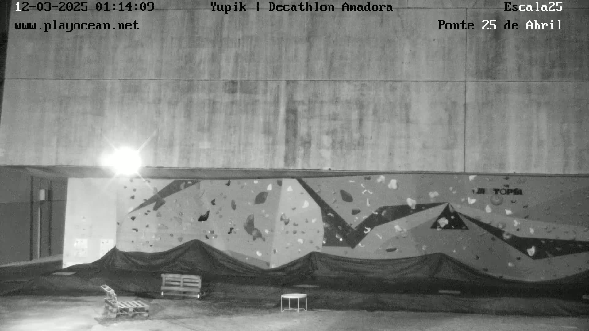 A nighttime, outdoor view of a climbing wall covered by a black tarp, with a small table and a pallet furniture set in the foreground.