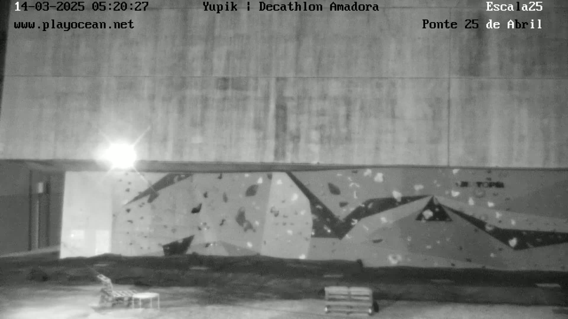 A nighttime, black-and-white image shows a large climbing wall under a bright light, with some furniture and a pallet visible in the foreground.