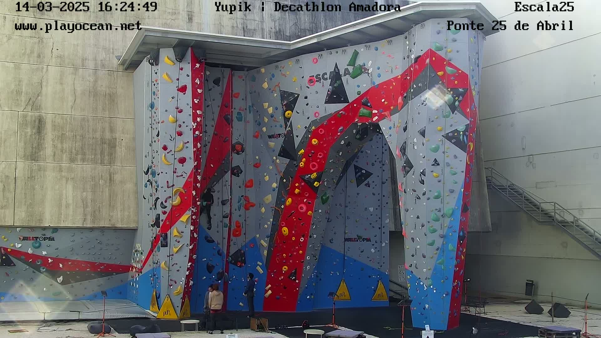 A large, colorful indoor climbing wall with several climbers present on a sunny day.
