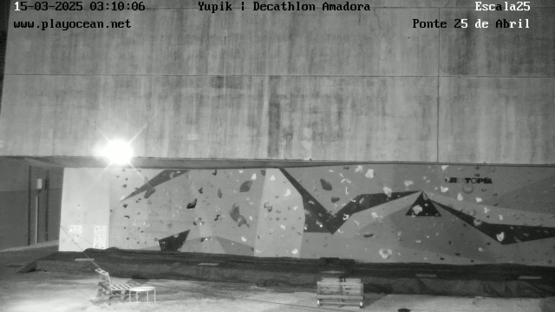 A nighttime black and white image shows an indoor climbing wall with various holds affixed to it, illuminated by a single overhead light, with a small amount of furniture visible in the foreground.