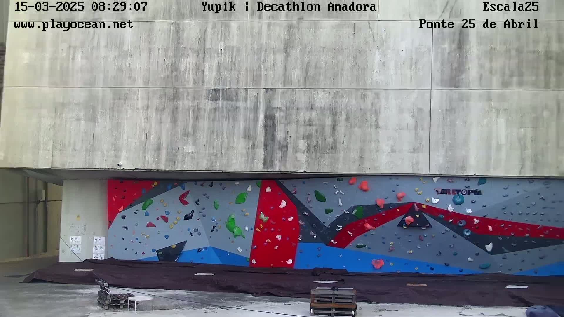 A large, outdoor climbing wall with various colored holds is situated against a concrete building under overcast skies.