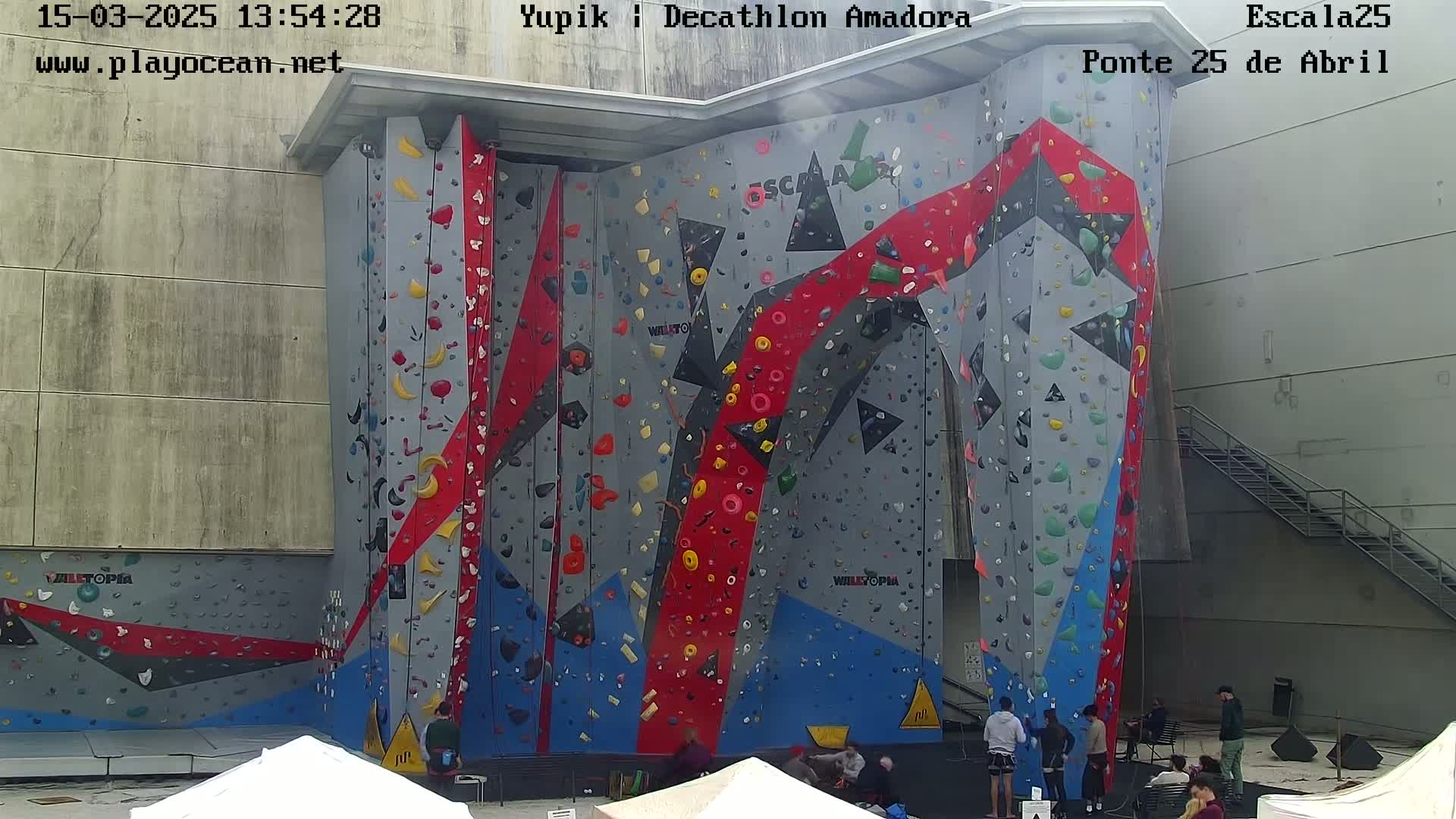 A large outdoor climbing wall with various colored holds is surrounded by people and covered tents, under a slightly hazy sky.