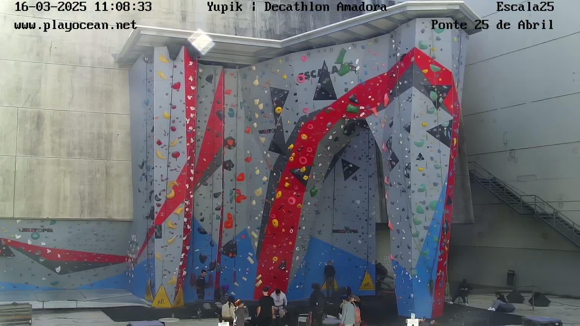 A large, colorful outdoor climbing wall stands against a building, with several people gathered at its base.