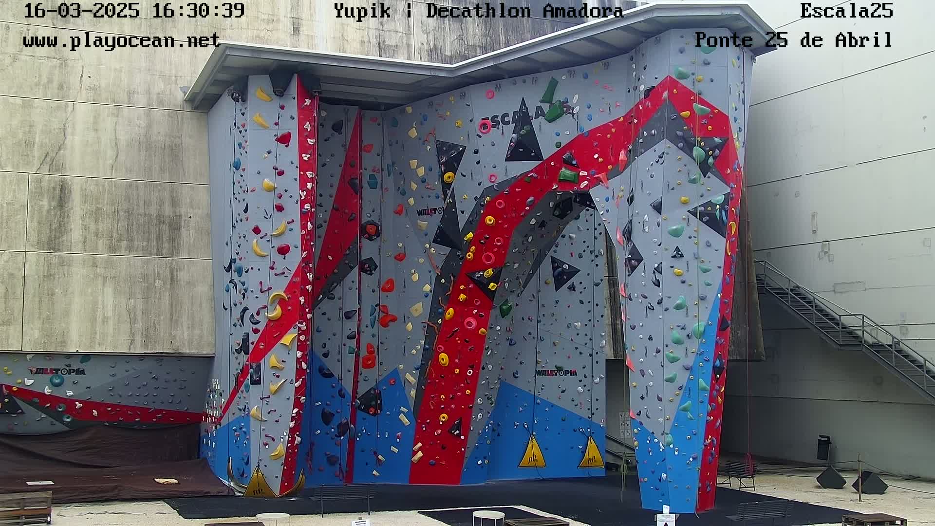 A large, multi-colored indoor climbing wall stands against a light gray building under an overcast sky.