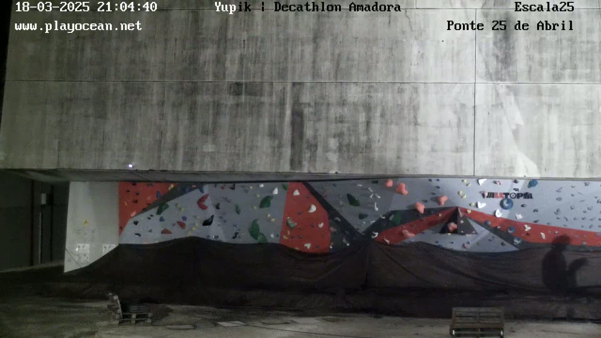 A nighttime view of an outdoor climbing wall with various colored holds, partially covered by a dark tarp.