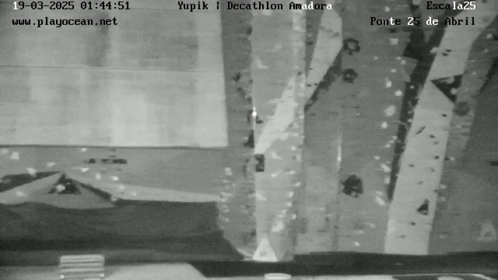 A grainy, black and white image shows a nighttime view of what appears to be a road or bridge, with vertical structures visible in the background.
