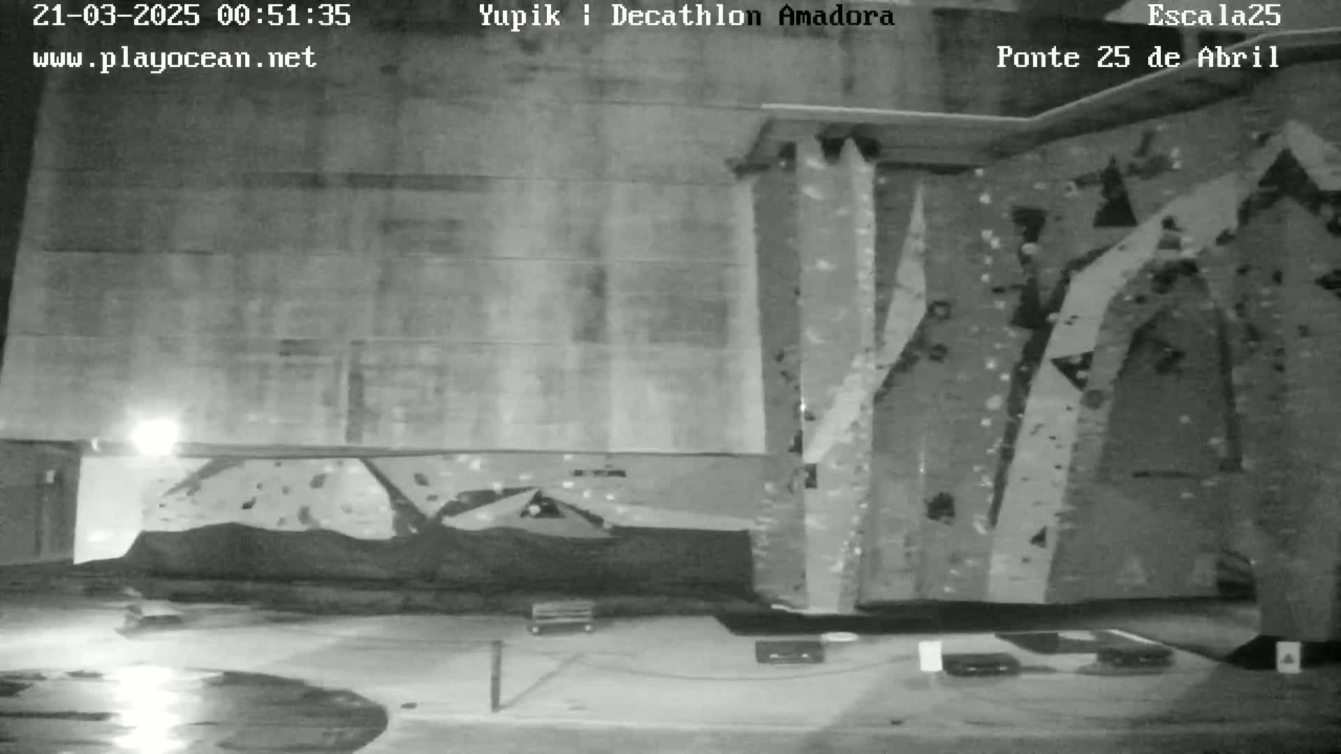 A nighttime, monochrome view of a large, textured climbing wall outdoors with a wet ground in front of it.