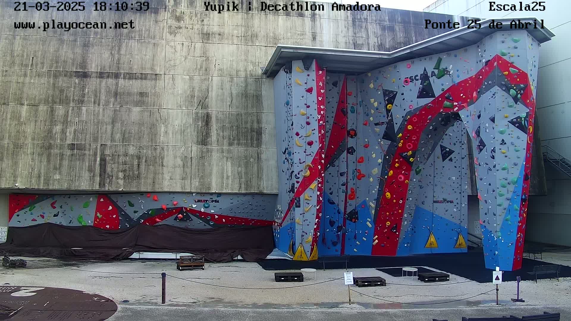 An outdoor climbing wall with red, blue, and gray sections stands against a larger gray building under a cloudy sky.