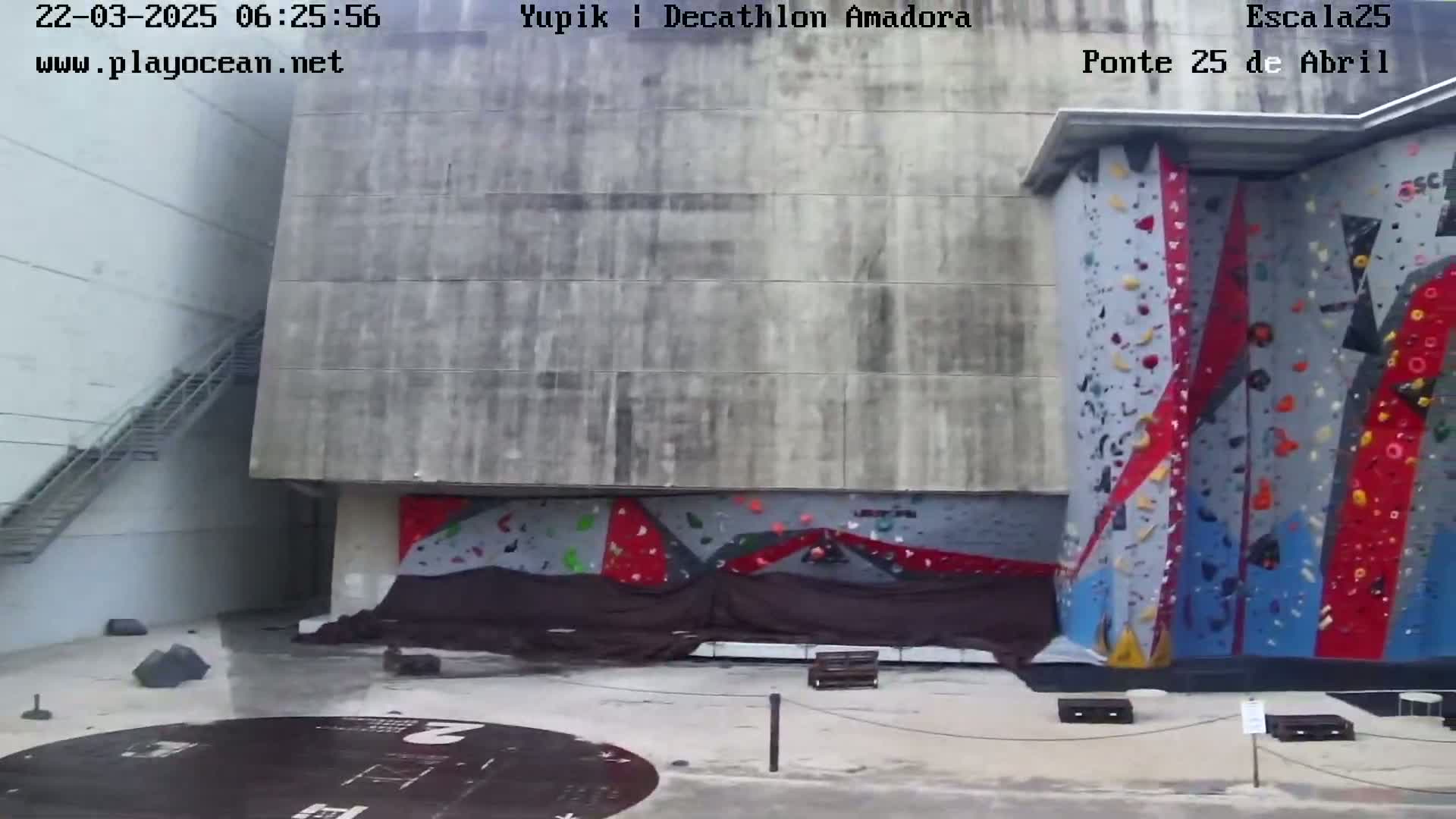 A large, outdoor climbing wall with colorful holds is partially covered by a dark tarp, situated next to a concrete building with a metal staircase.