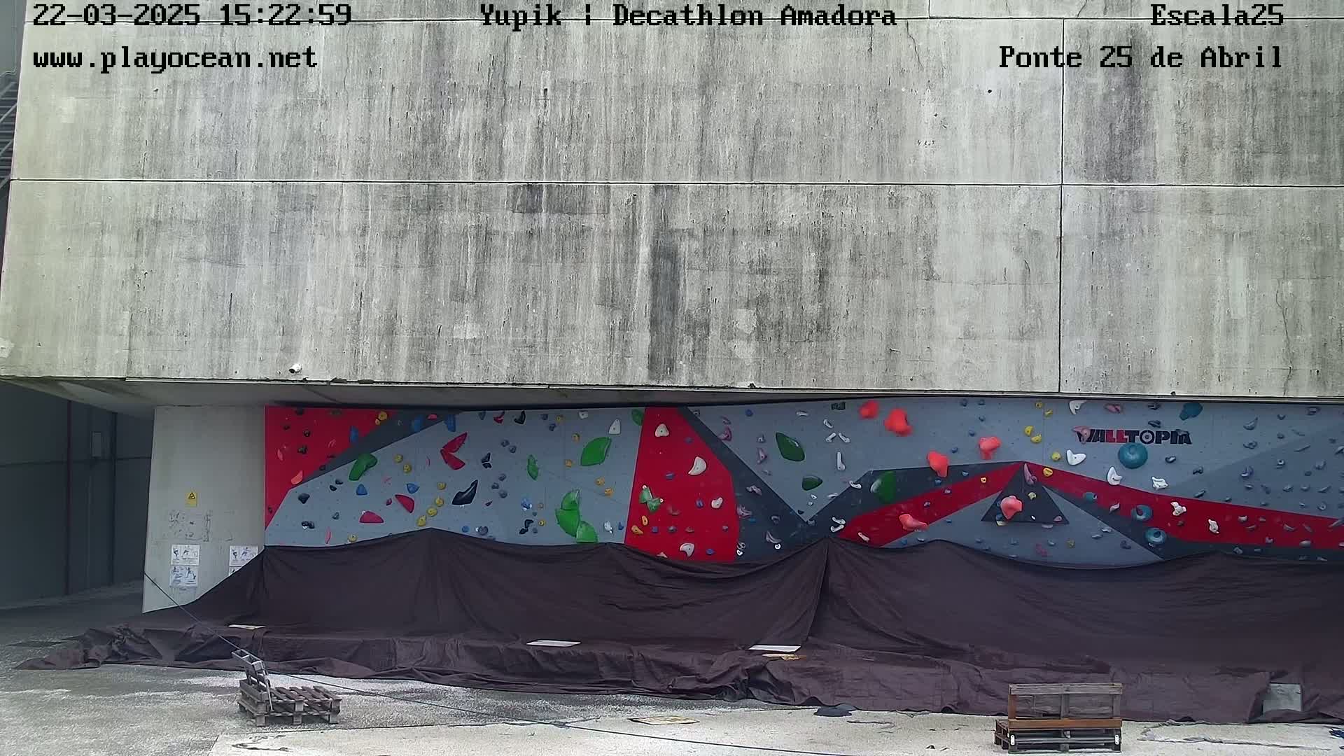 A large, multi-colored climbing wall is covered with a dark tarp, situated outdoors against a concrete building under an overcast sky.