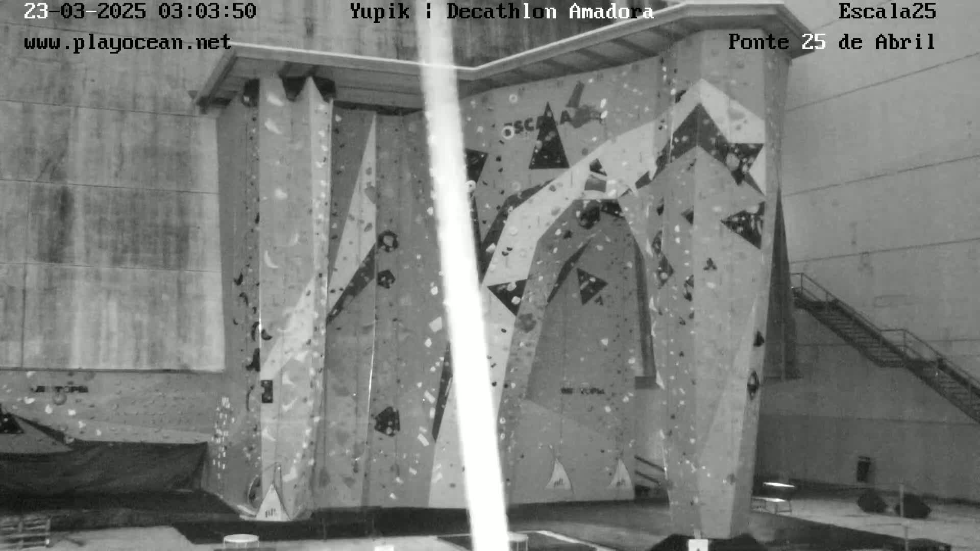 A grayscale image shows an indoor climbing wall with various holds and geometric patterns, in a large, empty room.