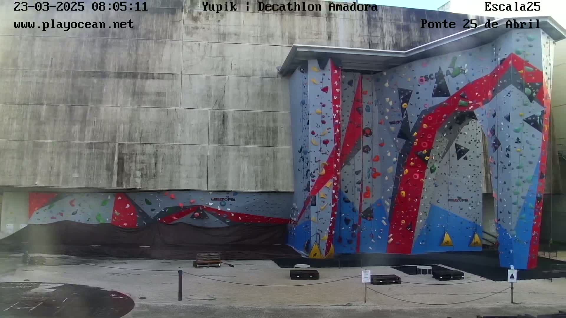 An outdoor view of a large climbing wall with various colored holds, set against a concrete building under a bright, sunny sky.