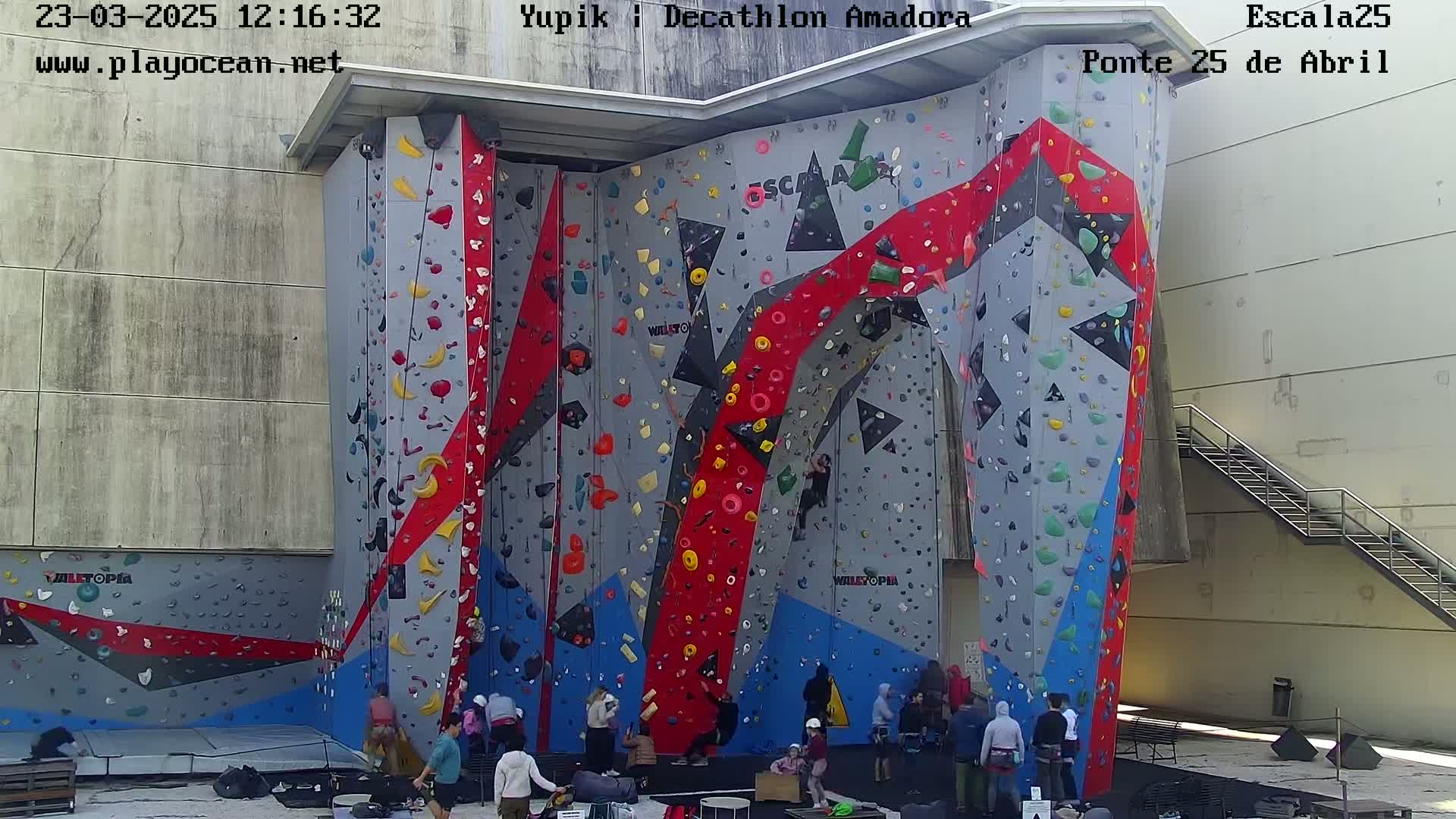 A large outdoor climbing wall with various colored holds, and several people are gathered around its base on a sunny day.