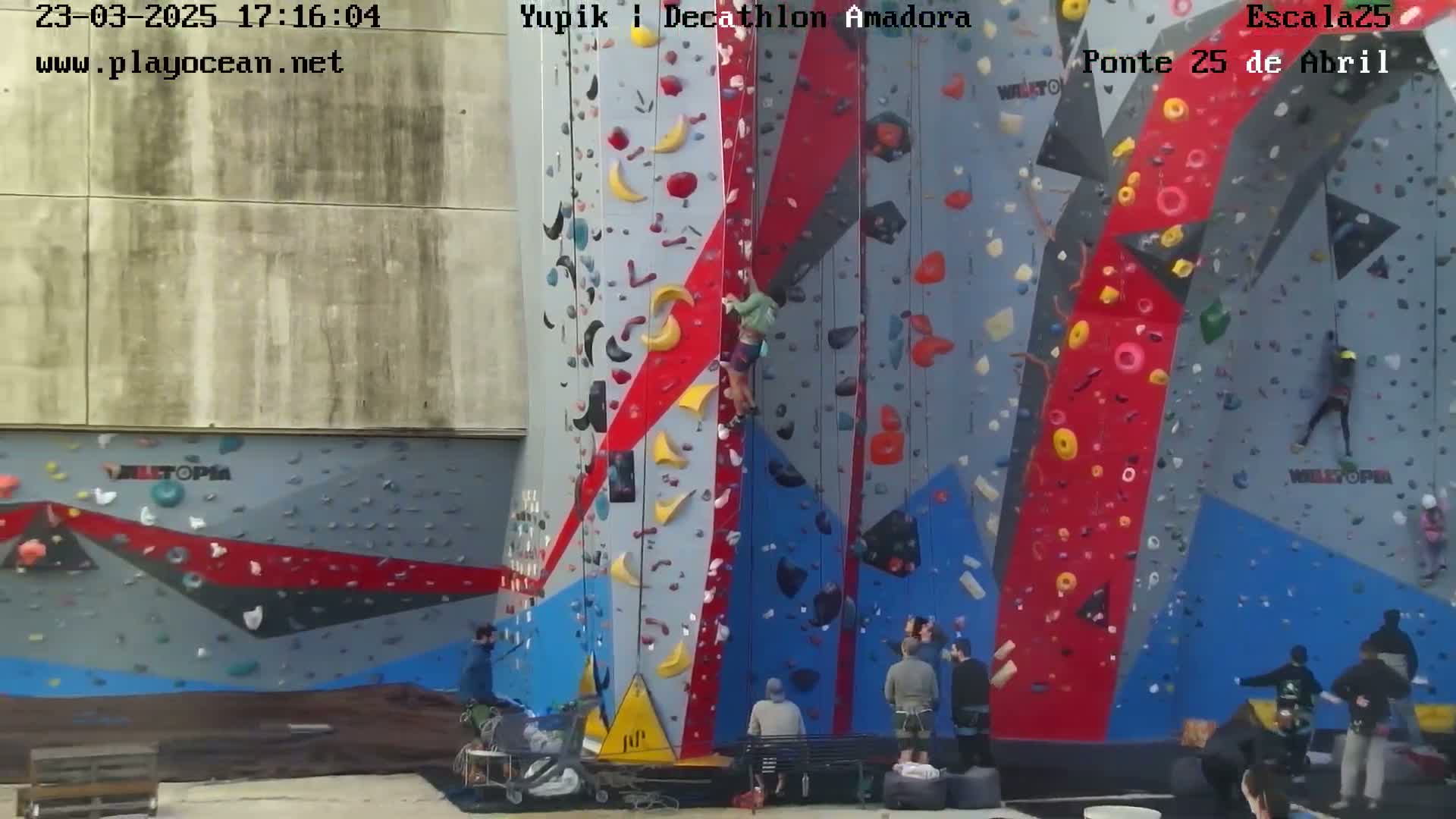 A large outdoor climbing wall with various colored holds and several people climbing or watching.