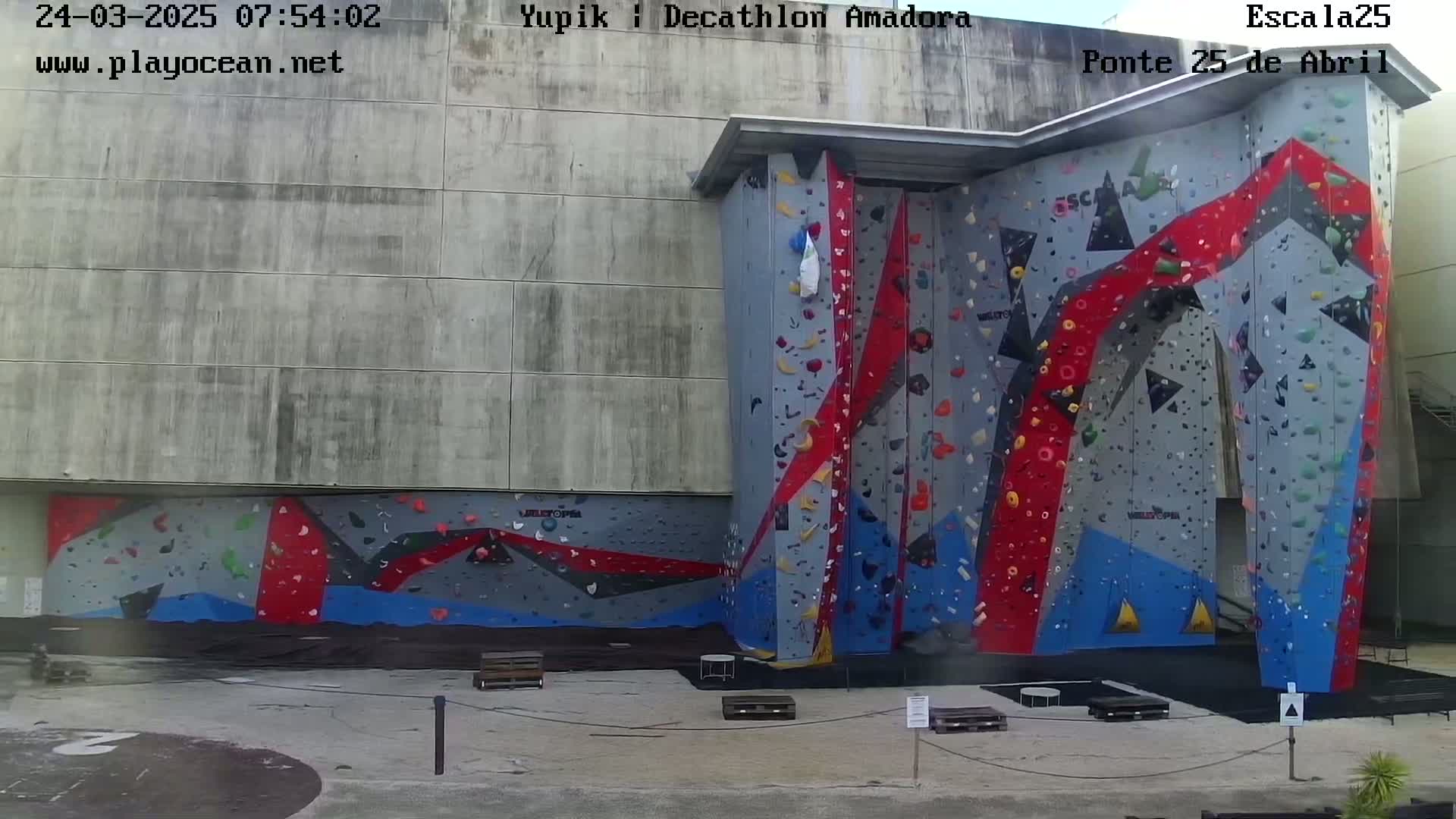 A large, multi-colored outdoor climbing wall is situated against a concrete building on a sunny day.