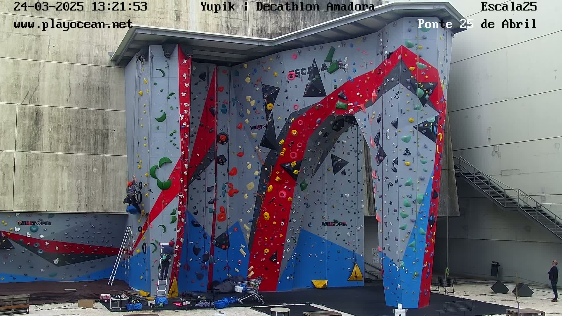 A large, multi-colored indoor climbing wall with several people climbing and others standing nearby.