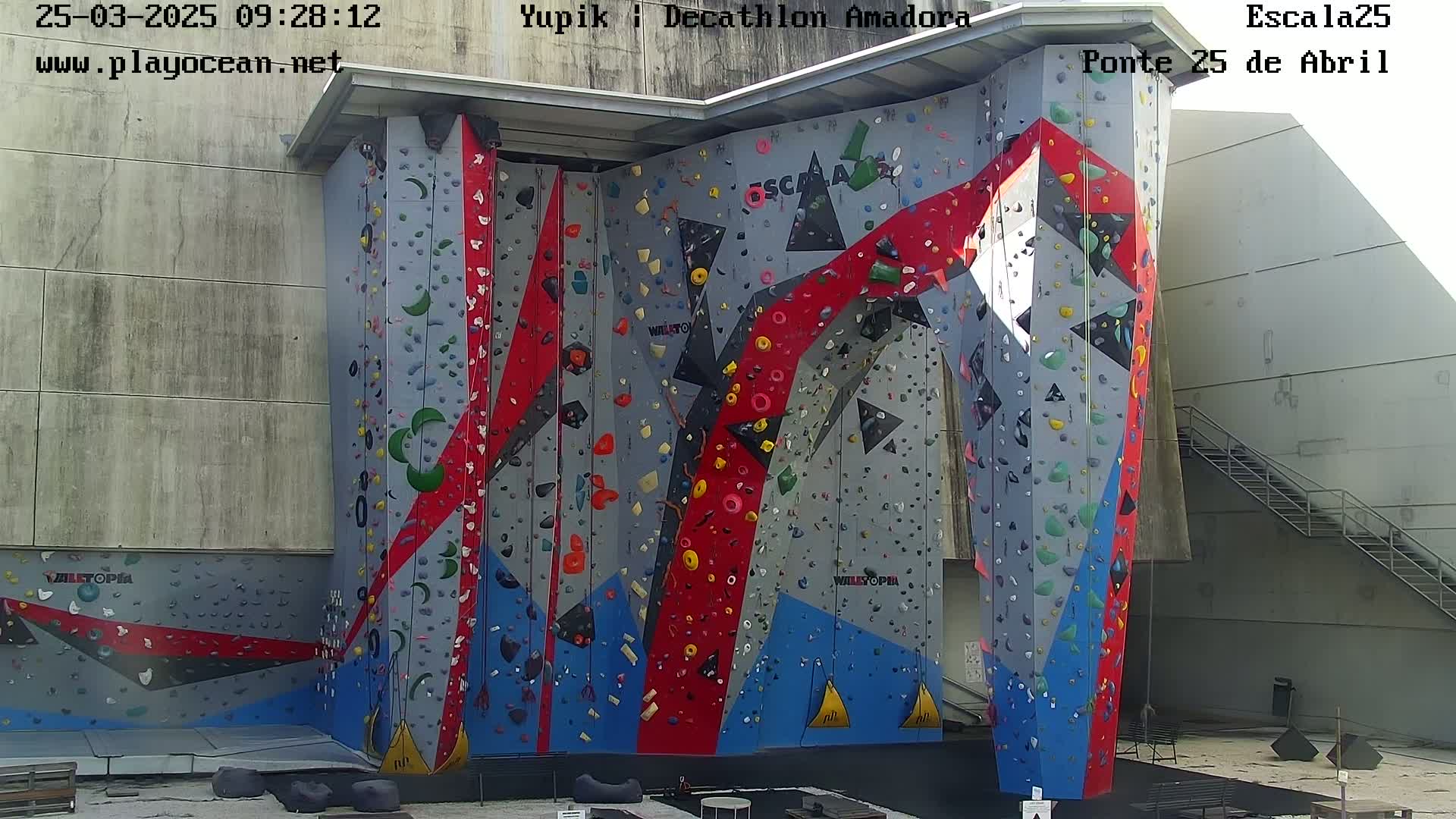 A large, colorful indoor climbing wall with various holds stands against a light gray building on a mostly sunny day.