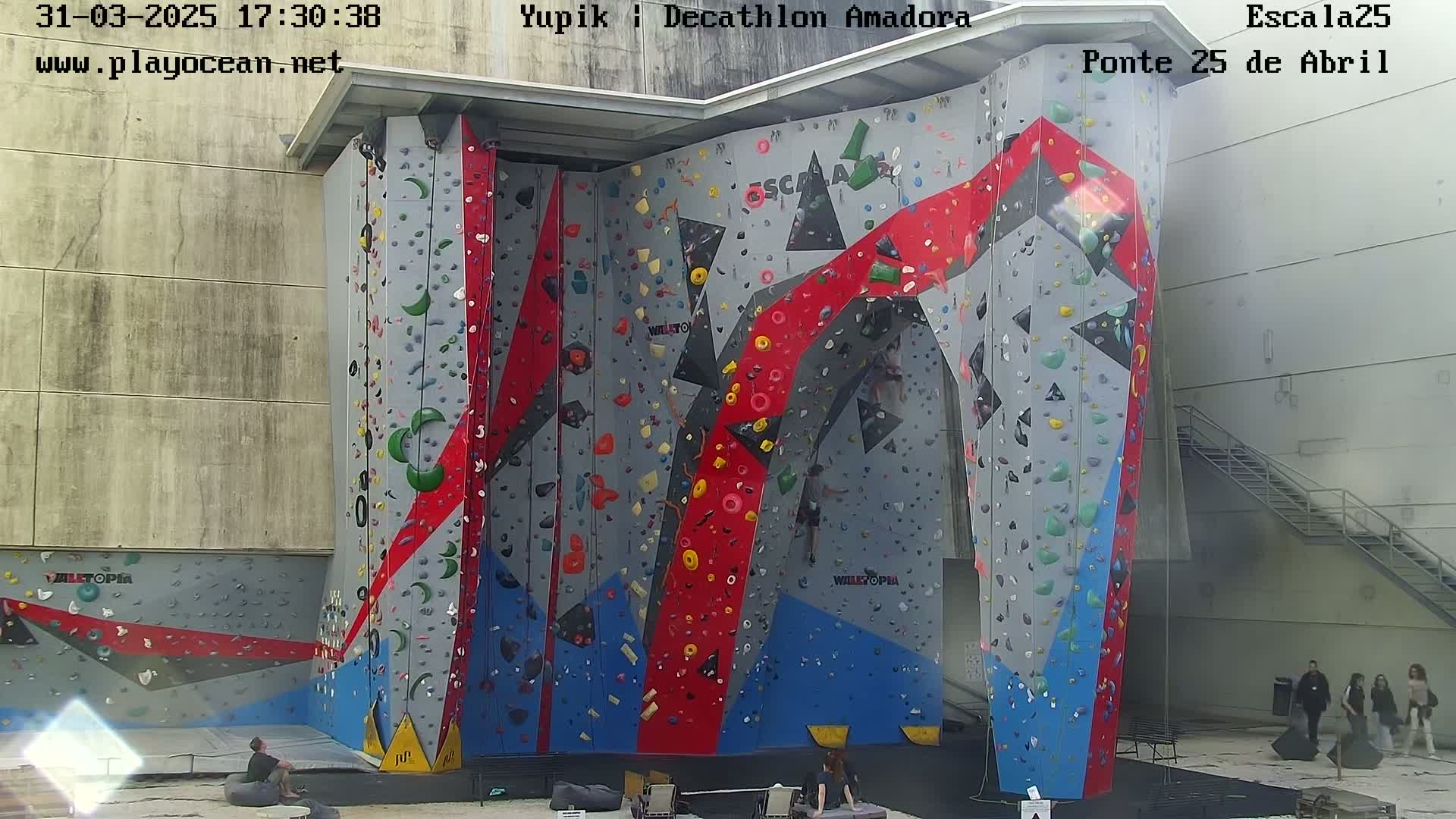 A large indoor climbing wall with various colored holds is situated in a building, next to an outdoor staircase.