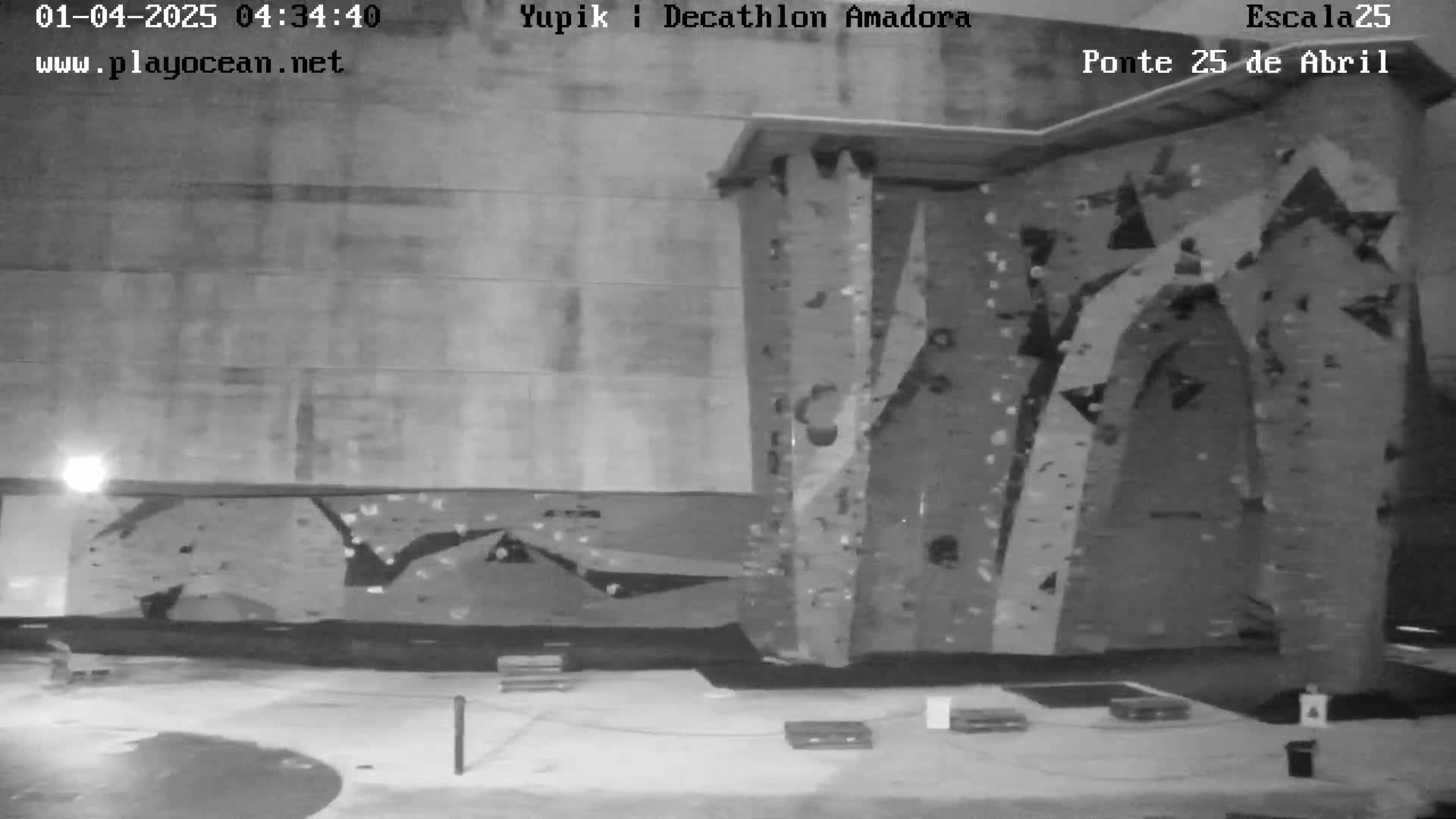 A nighttime, outdoor view of a large, textured climbing wall stands against a building, with a mostly empty area in front.