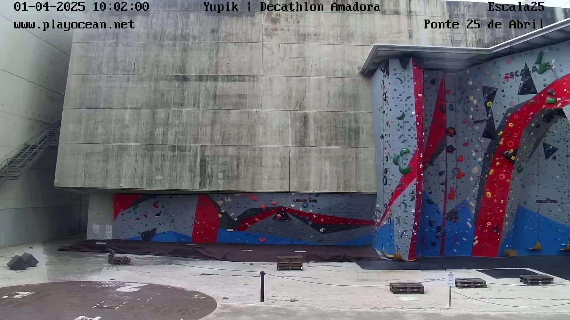 A large outdoor climbing wall with red, blue, and gray holds is situated against a concrete building.