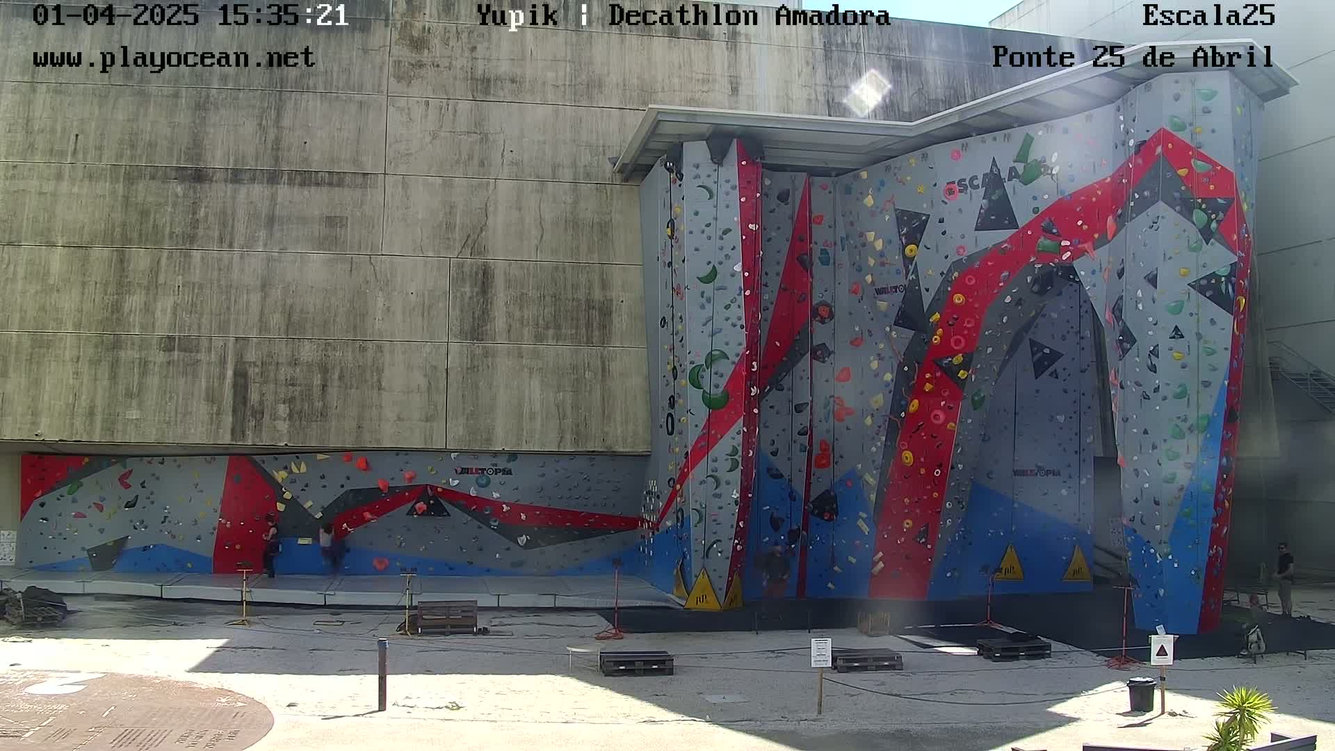 A large outdoor climbing wall with various colored holds is situated against a building on a sunny day.