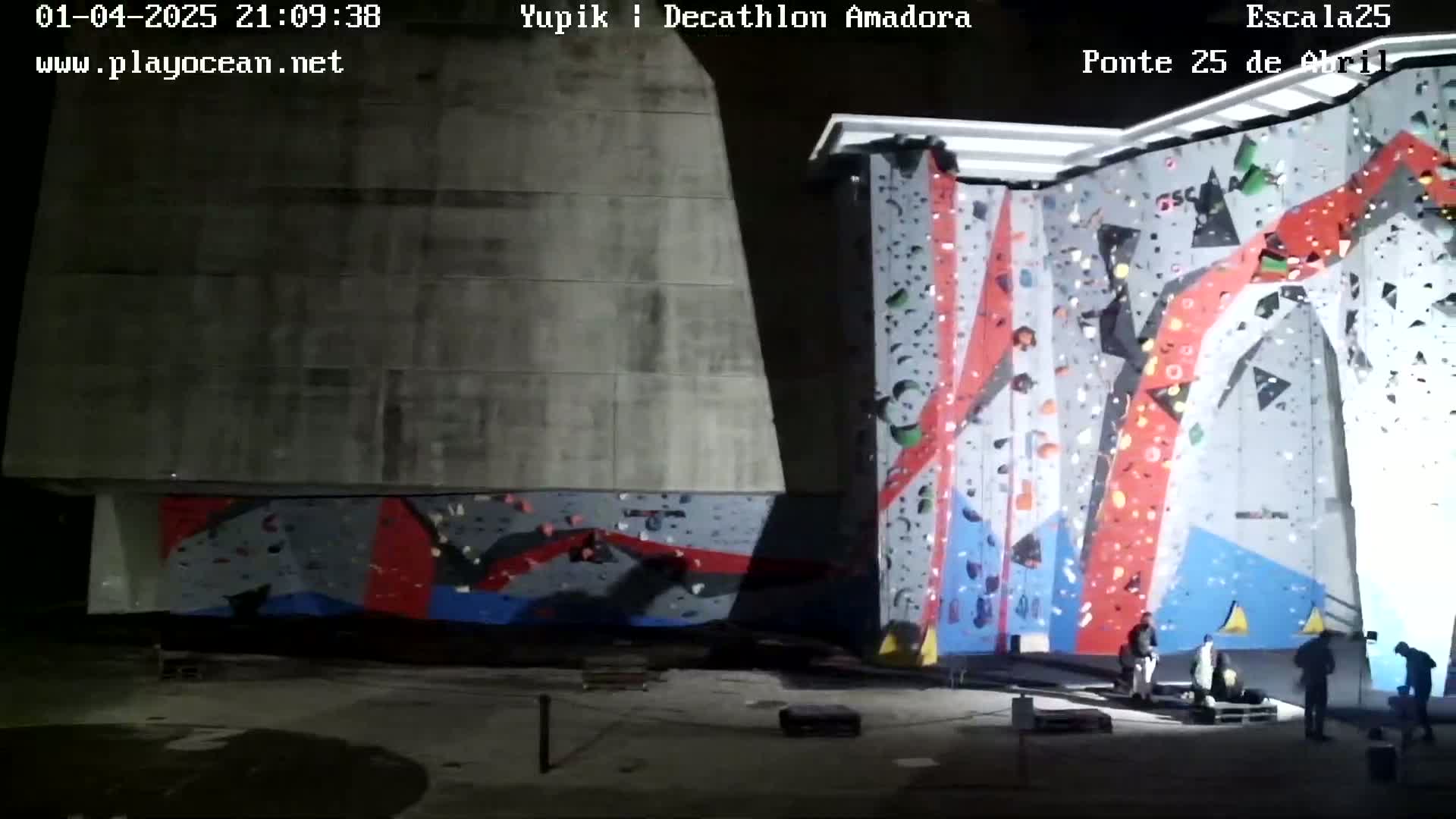 At night, several people stand near large, brightly colored indoor climbing walls.