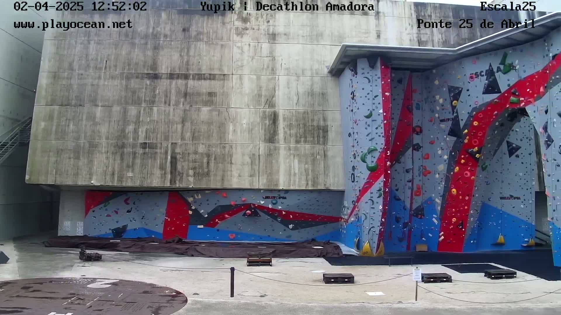 A large outdoor climbing wall with red, blue, and gray holds is situated against a concrete building under overcast skies.