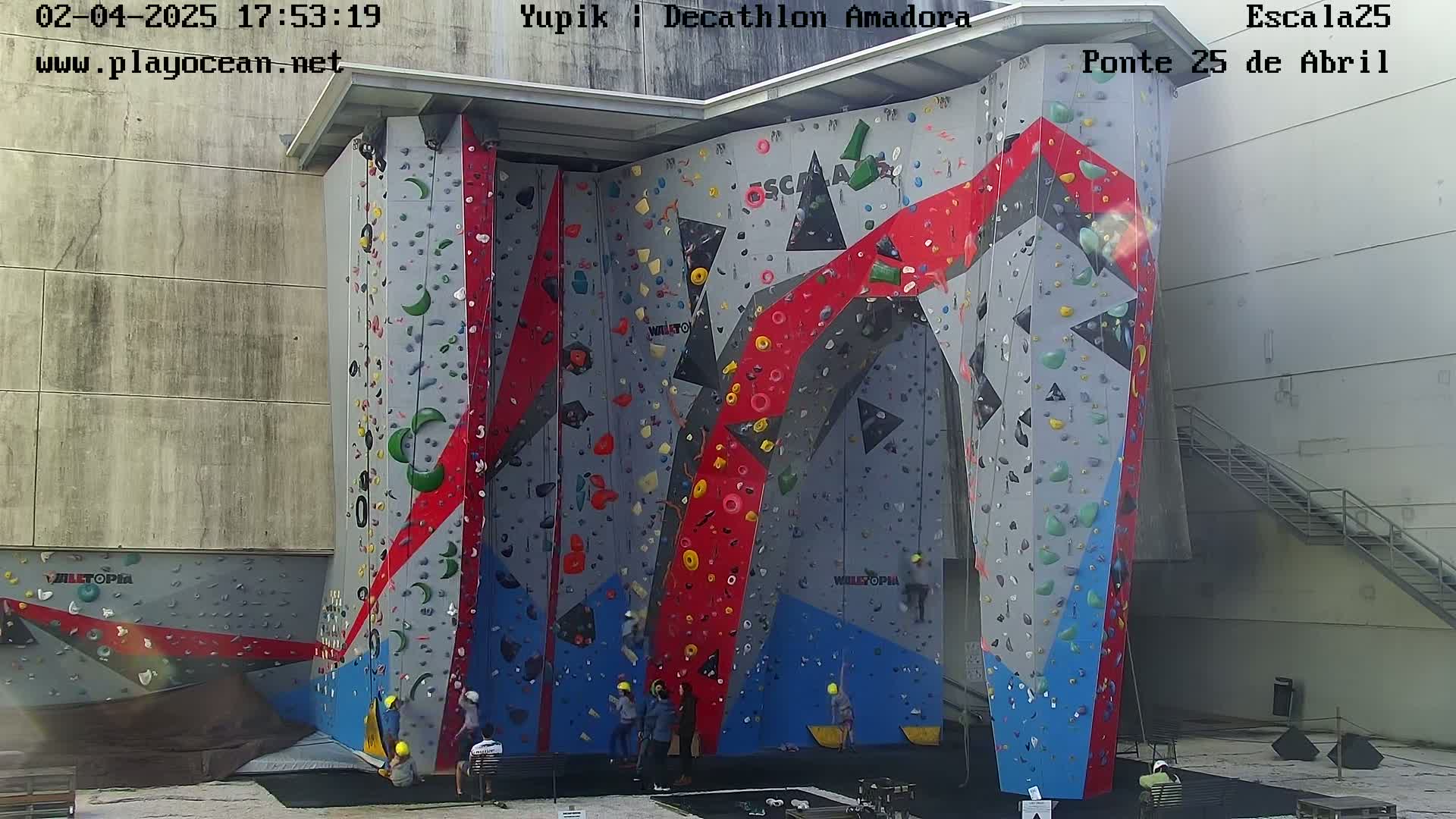 A large, colorful outdoor climbing wall with several people near its base, under a partly sunny sky.