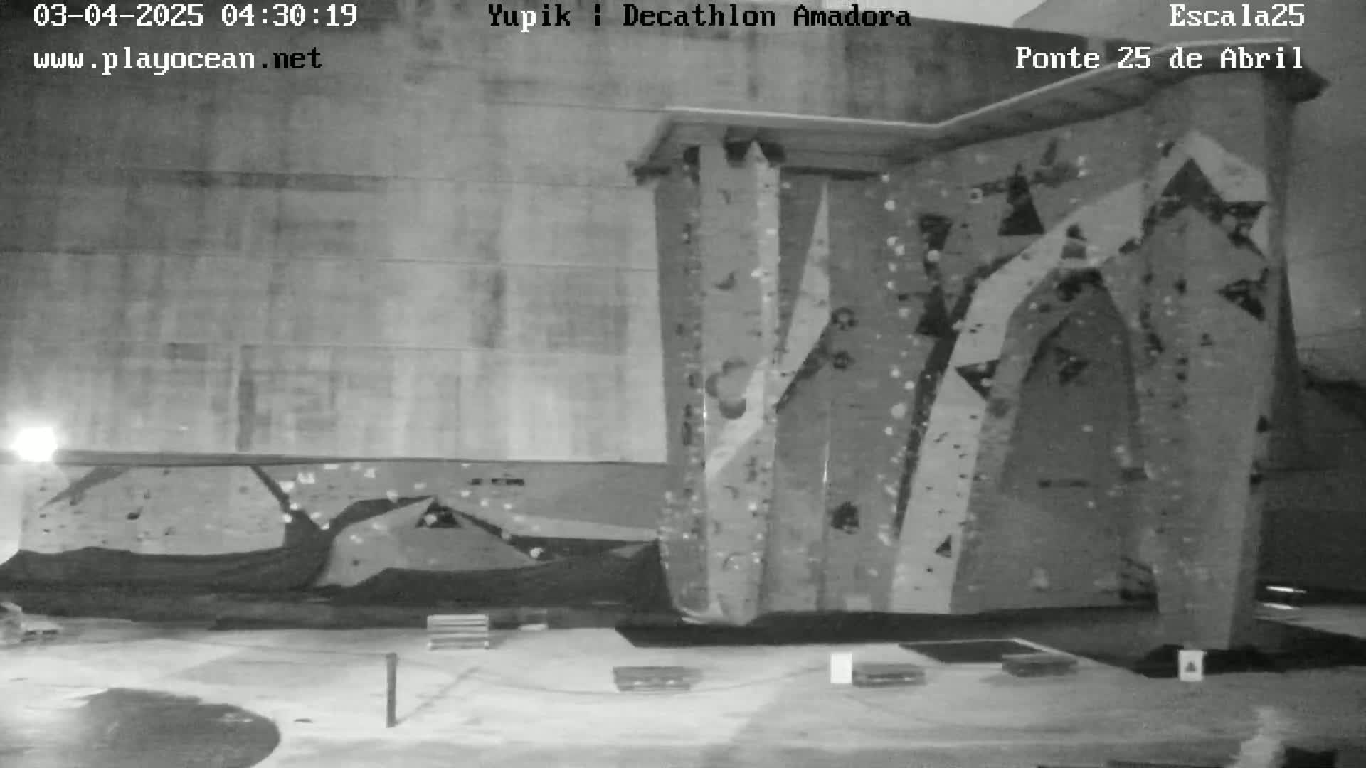 A nighttime, black and white image shows a large outdoor climbing wall next to a building under dim lighting.