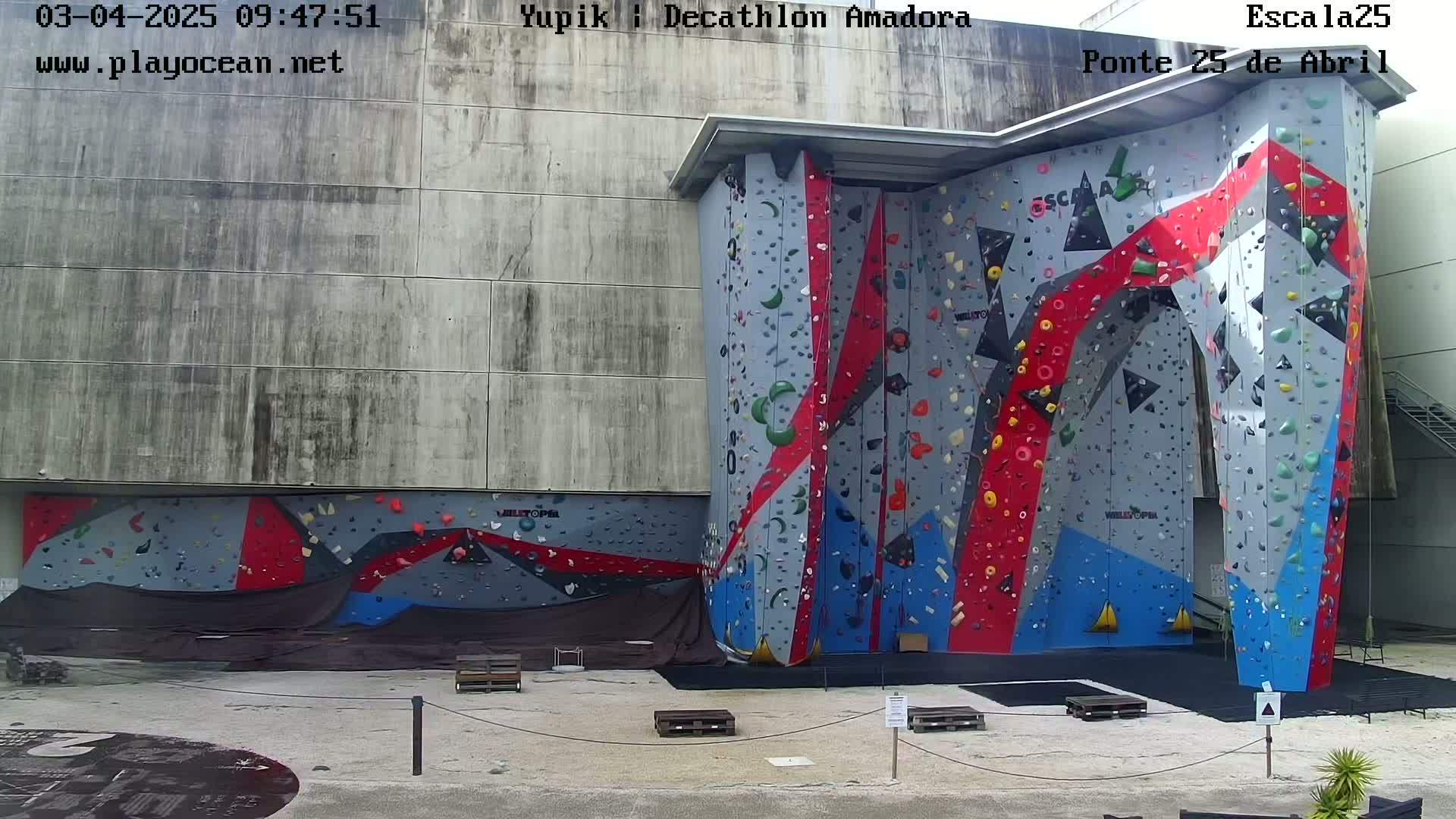 A large, multicolored climbing wall is situated outdoors on a cement surface, under a partly cloudy sky.