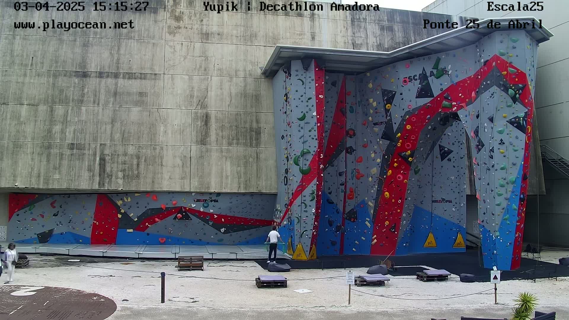 A large outdoor climbing wall with red, blue, and gray sections, and two people standing near it on a mostly clear day.