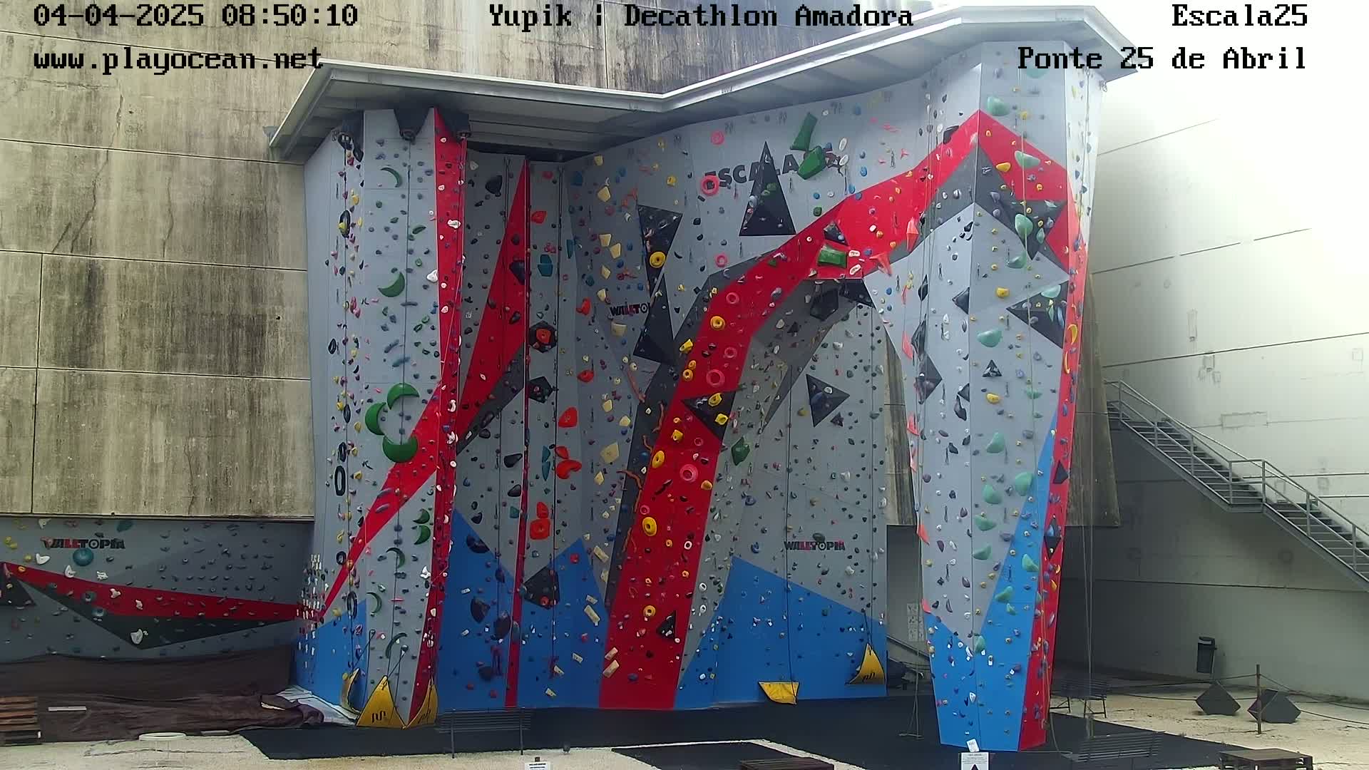 An outdoor climbing wall with various colored holds and sections is situated next to a light gray building with a metal staircase.