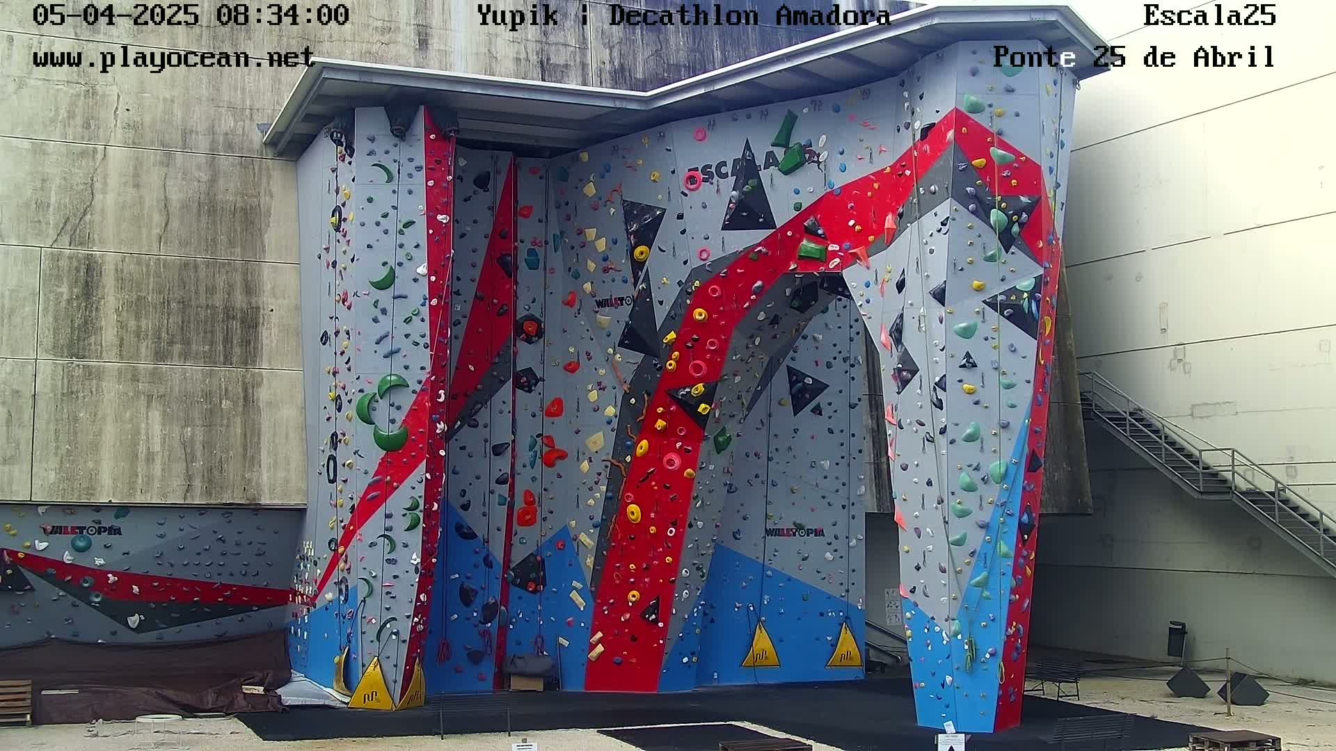 A large, colorful indoor climbing wall stands against a light gray building on a partly sunny day.