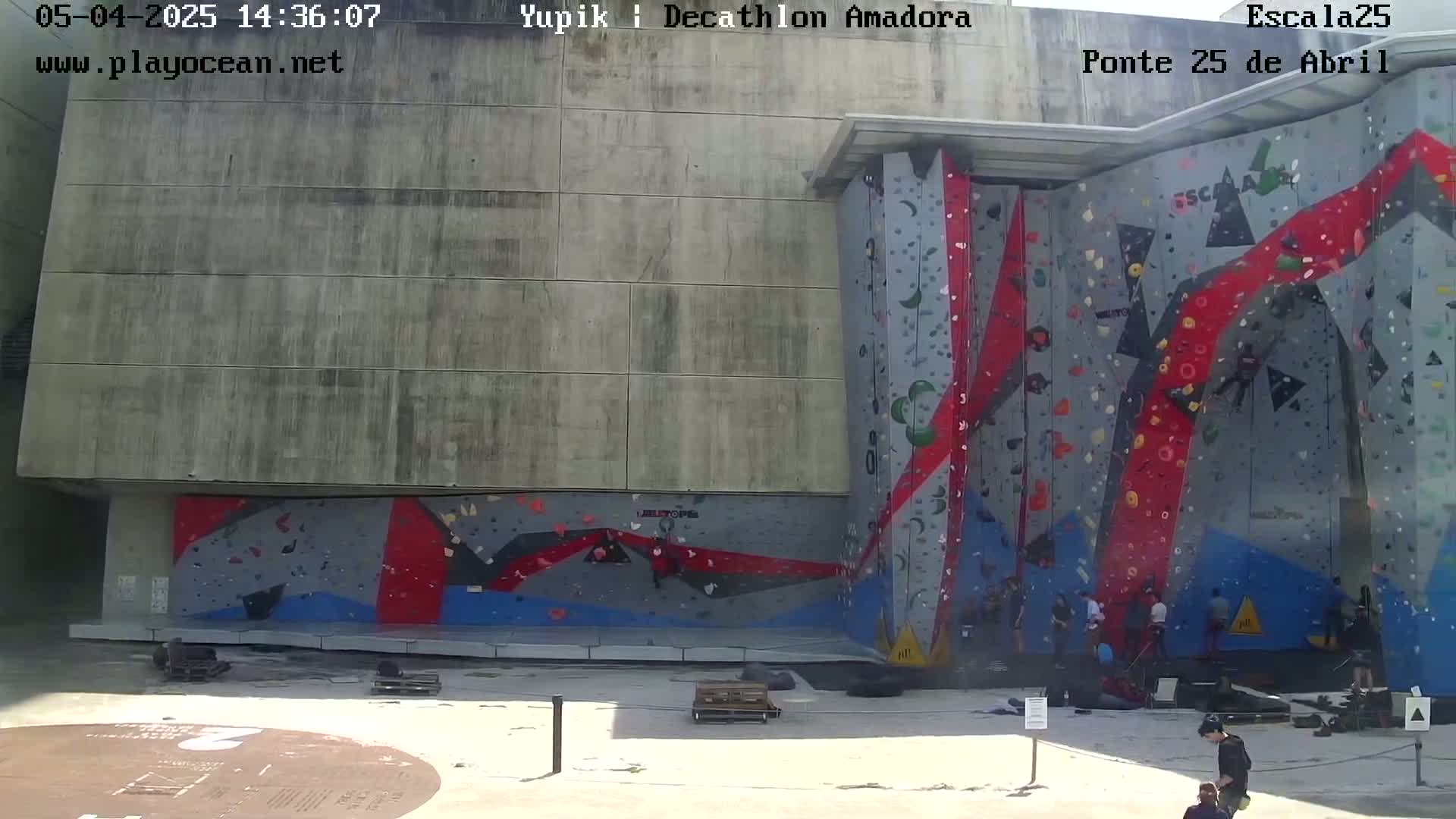 A large outdoor climbing wall with various colored holds, several people climbing and watching, and a mostly sunny day.
