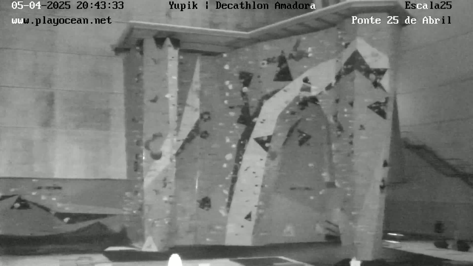 A grayscale indoor image shows a large climbing wall with various holds and routes.