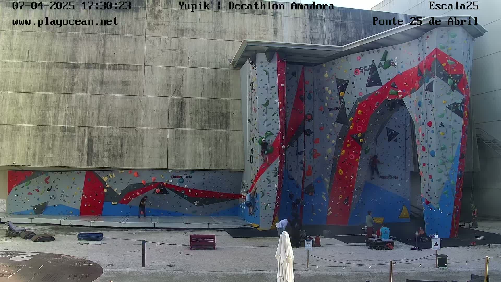 An outdoor climbing wall with several people climbing and others watching, under a mostly sunny sky.