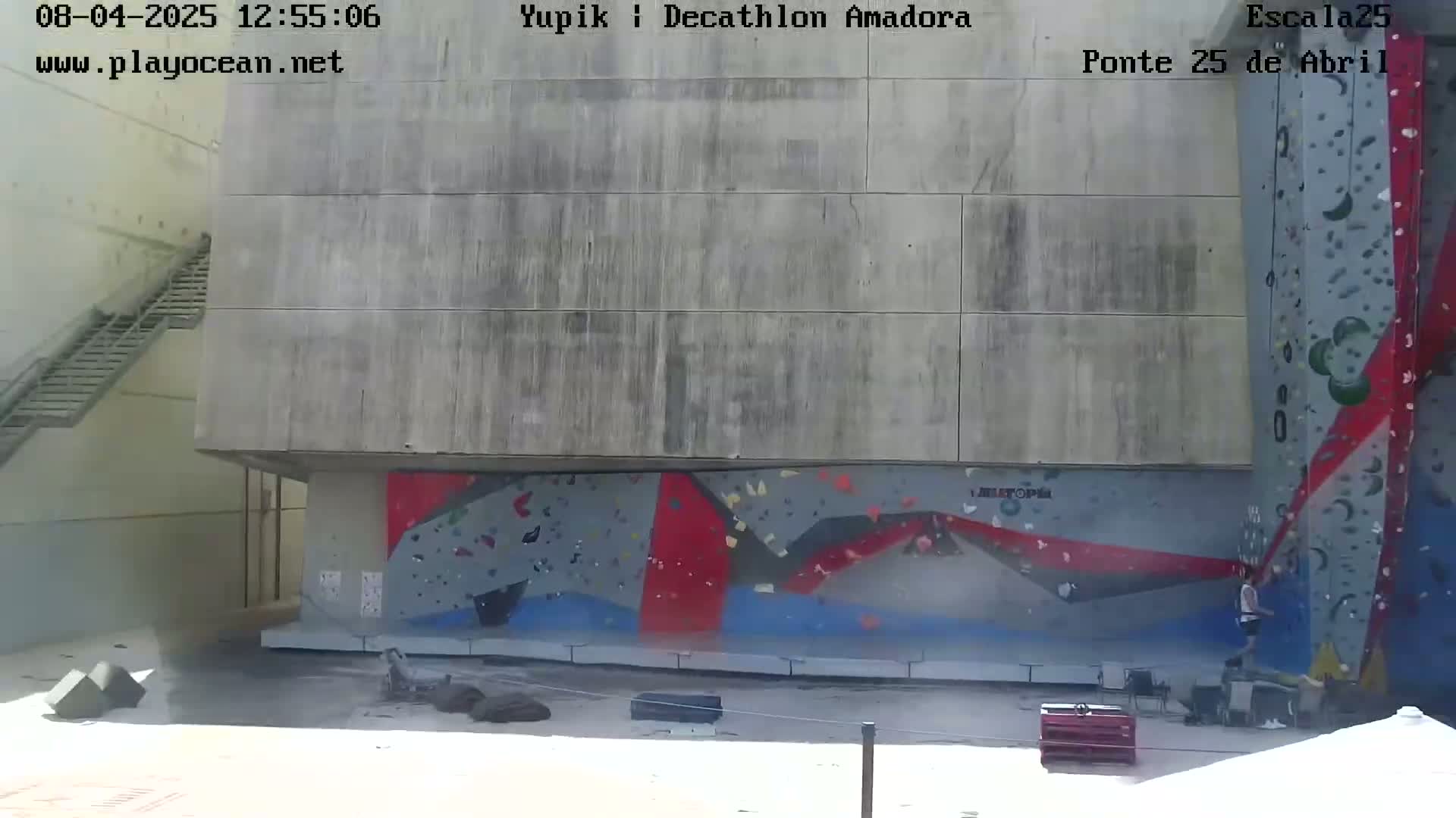 A person climbs an outdoor climbing wall painted with geometric shapes on a mostly sunny day.