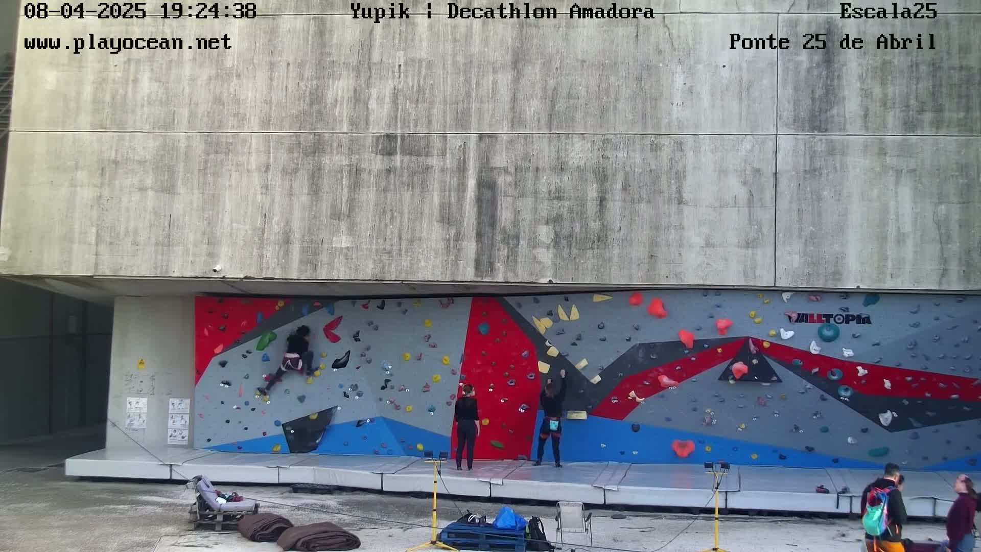 Several people are rock climbing on an outdoor wall, with some people observing from below on a concrete area.