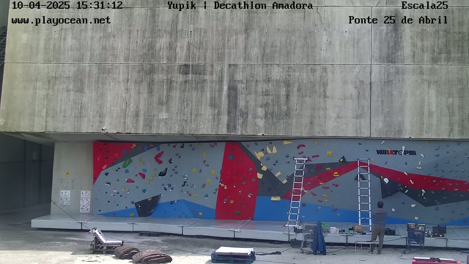A large, outdoor climbing wall with various colored sections is set against a concrete building under a bright, sunny sky, and two people stand near it.