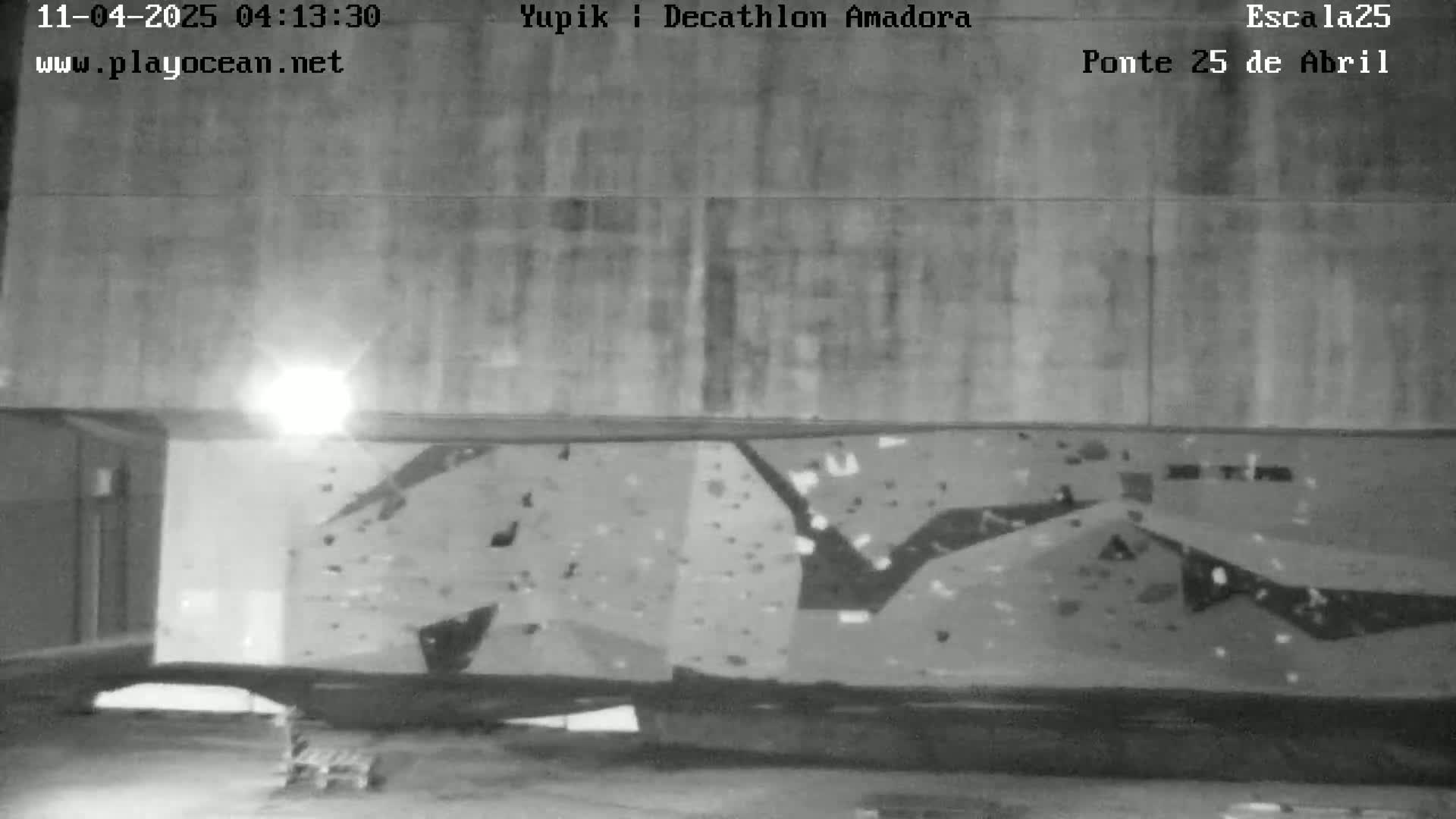 A grayscale night image shows a large, textured climbing wall affixed to a building's exterior under artificial light.
